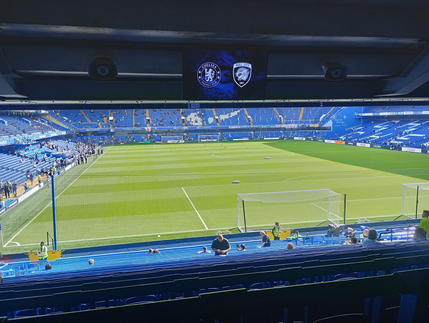 A football stadium with a well-maintained pitch and a large screen displaying the logos of Chelsea FC and their opponent, with a few spectators seated.