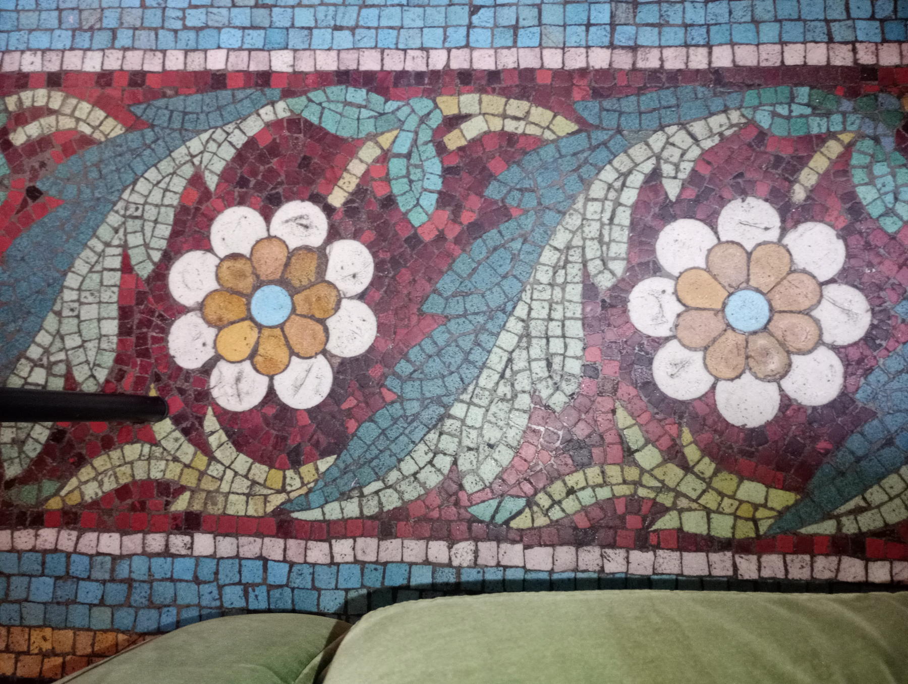 A vibrant mosaic floor features colorful floral patterns with white petals and blue centers surrounded by green leaves against a red and blue background.