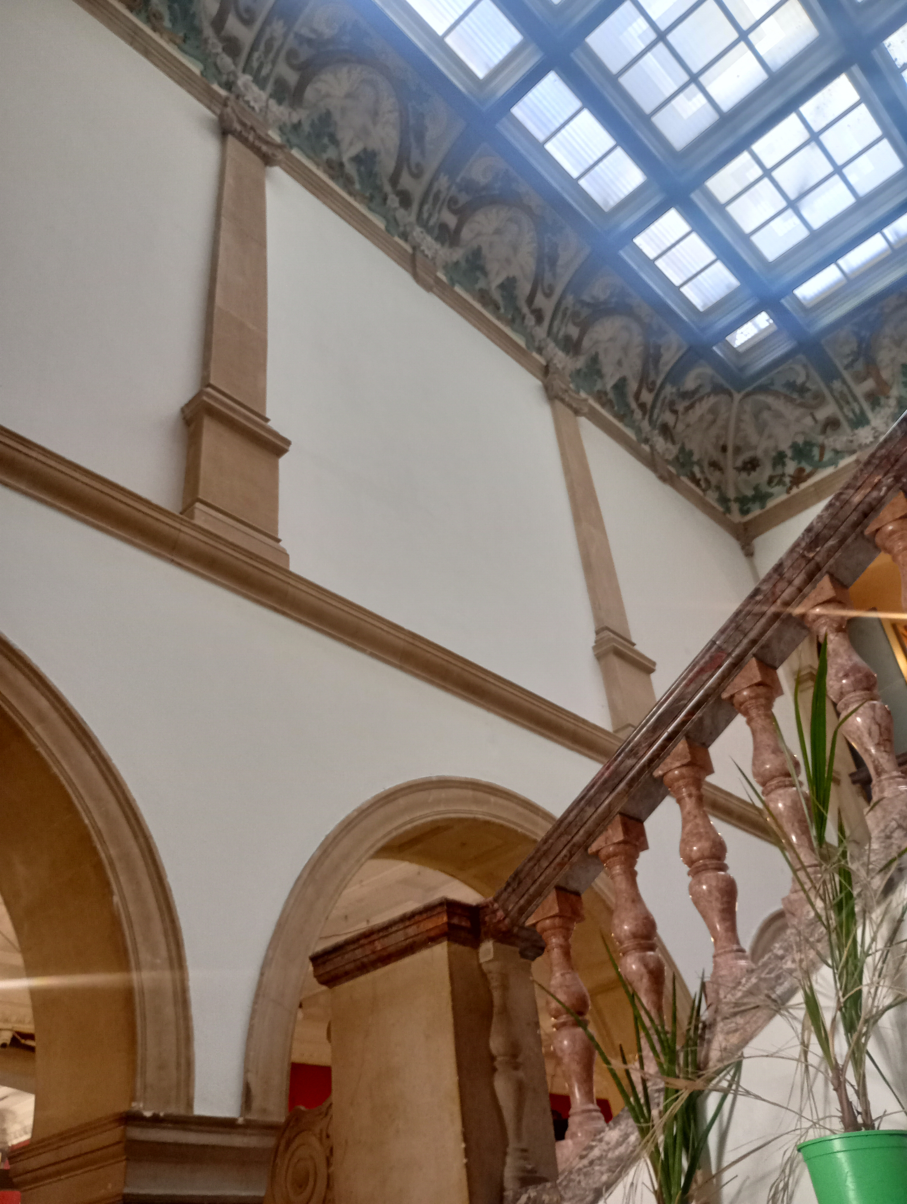 A decorative indoor staircase with stone balusters leads upward under a skylight, framed by arched doorways and a painted frieze near the ceiling.