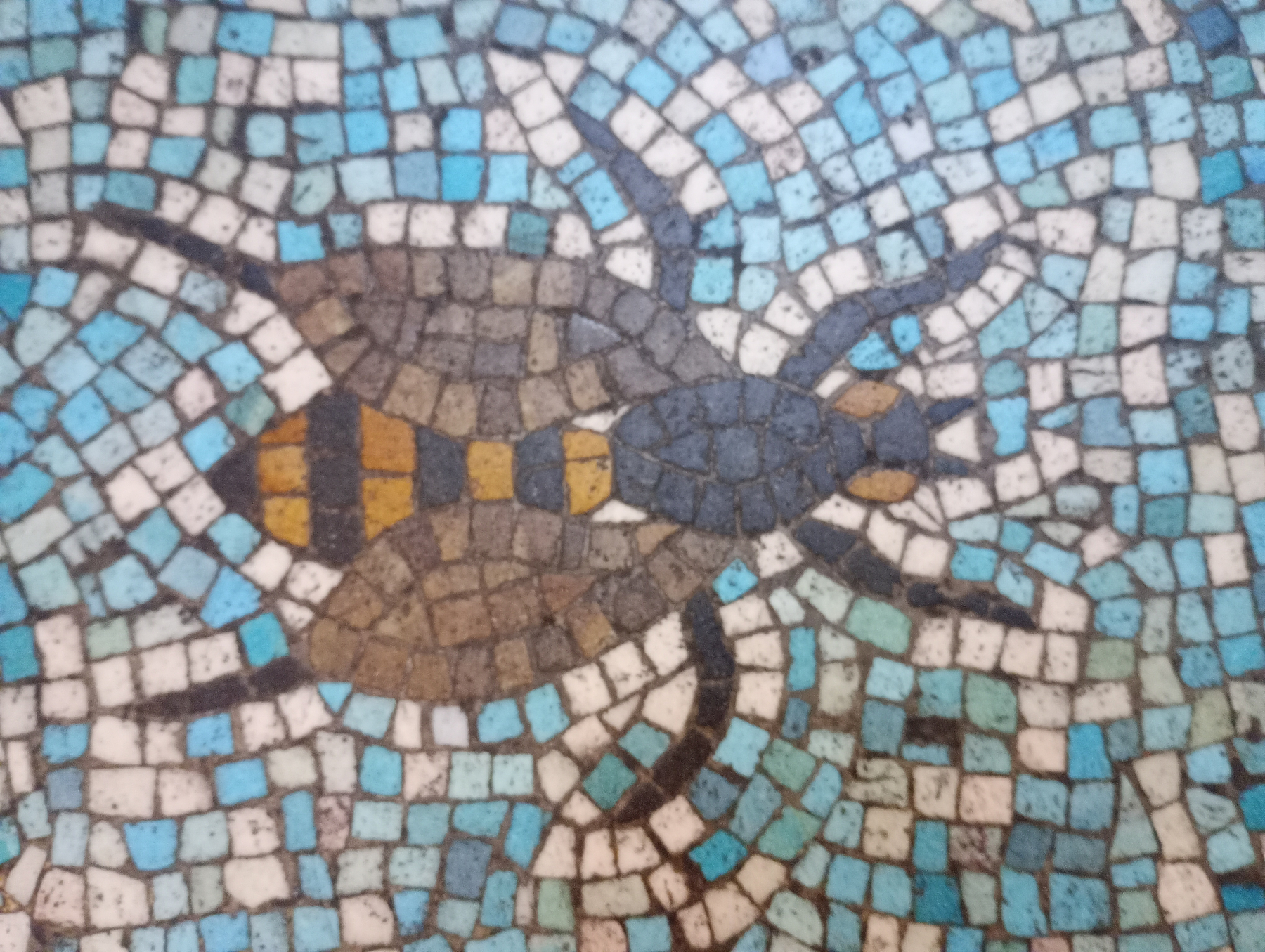 A mosaic artwork depicts a bee with a blue head, brown and yellow striped body, and wings, set against a background of blue and white tiles.