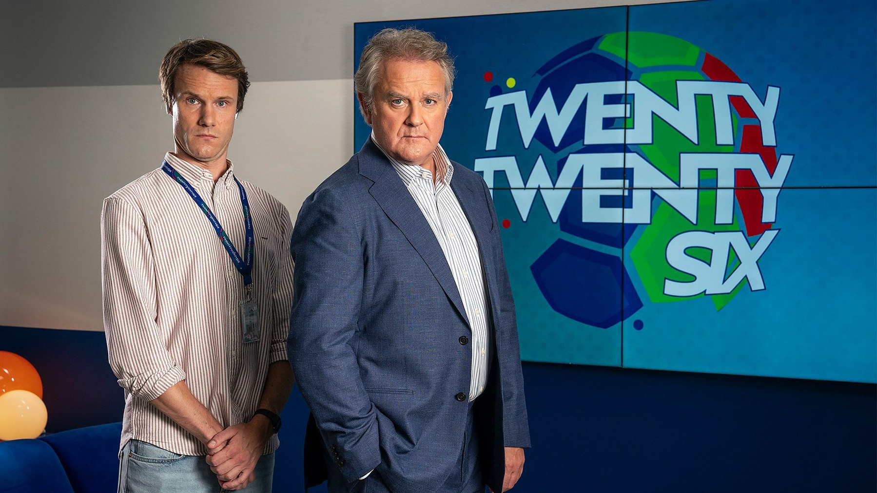 Two men stand in front of a screen displaying the text TWENTY TWENTY SIX, with one wearing a casual striped shirt and the other in a suit.