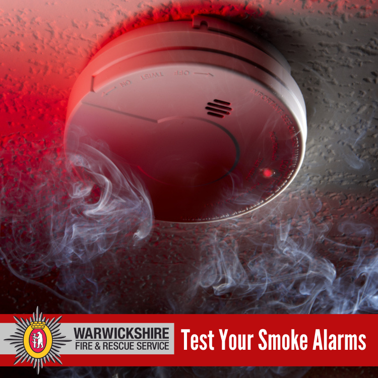 A smoke alarm mounted on a ceiling is surrounded by smoke with a red alert light, accompanied by a warning to test smoke alarms from the Warwickshire Fire & Rescue Service.