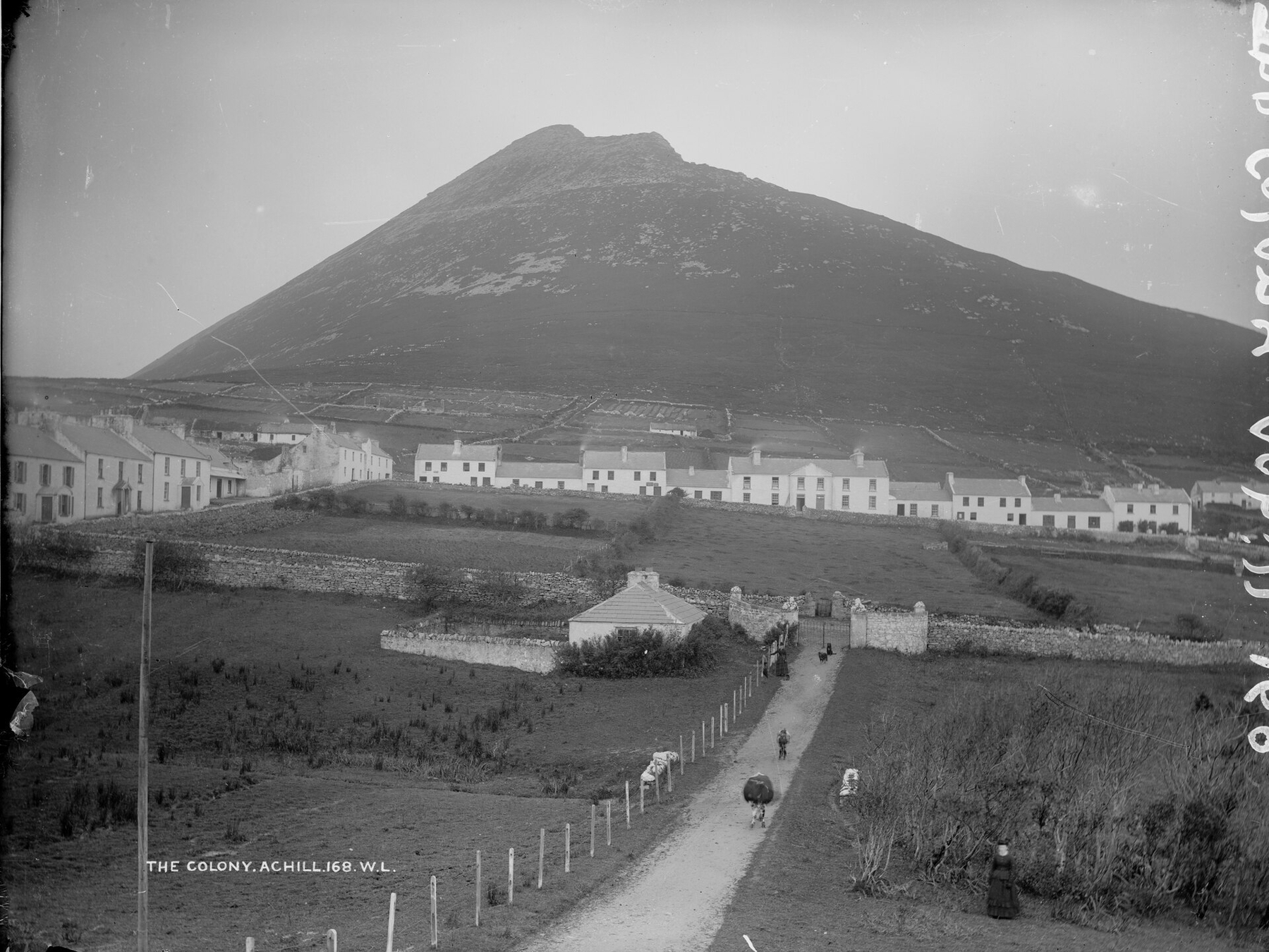 A rural settlement with white houses is situated at the base of a large, rocky hill with a pathway leading toward it.