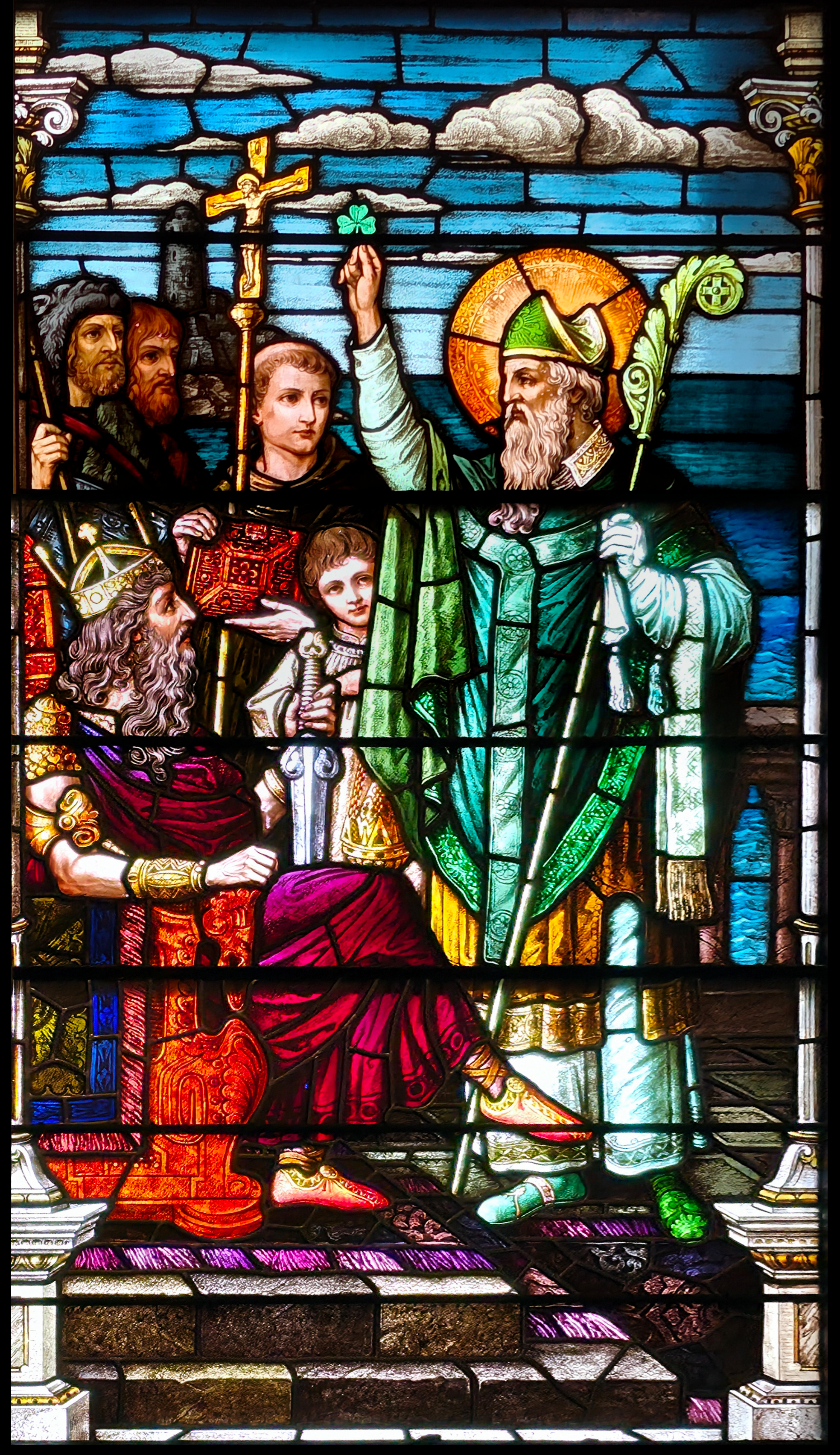 A stained glass window depicts a religious figure holding a shamrock and speaking to a seated king, surrounded by attendants.