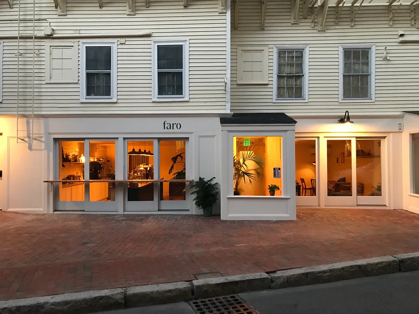 A warmly lit café front with large windows on a traditional two-story building, featuring a clean, modern design and the name 'faro' above the windows. The exterior is painted white with a brick pavement, and it's twilight outside.