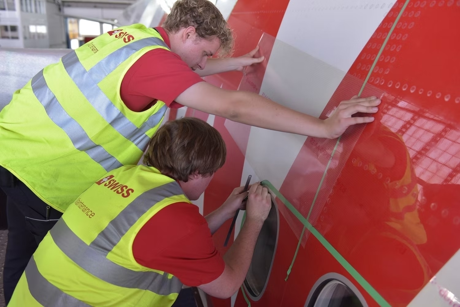 Two workers apply a shark skin-like coating, AeroSHARK, to an airplane’s red and white exterior, carefully working around a window with precision tools.