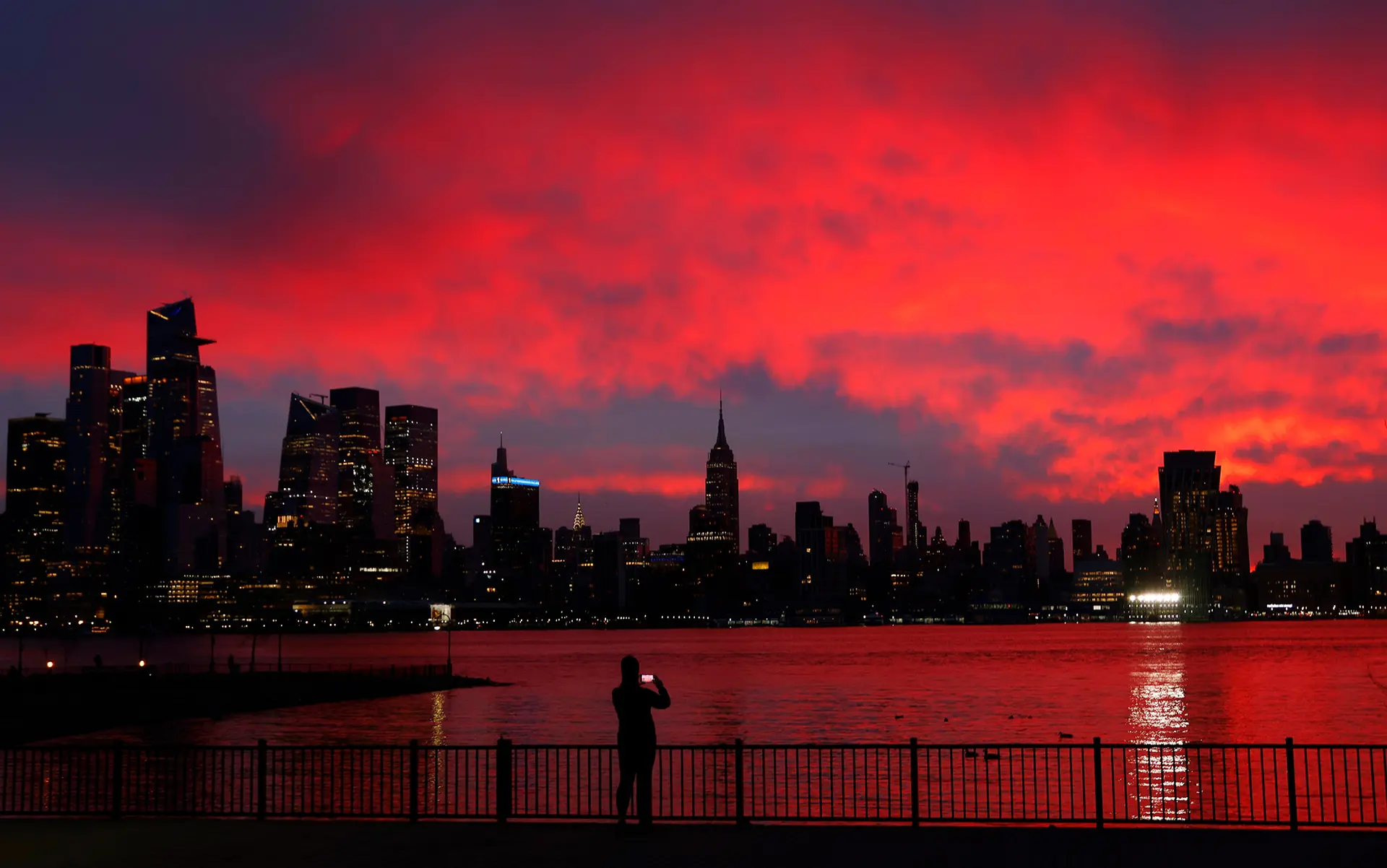 A silhouette of a person taking a photo of a city skyline against a vibrant red sunset reflected in the water.