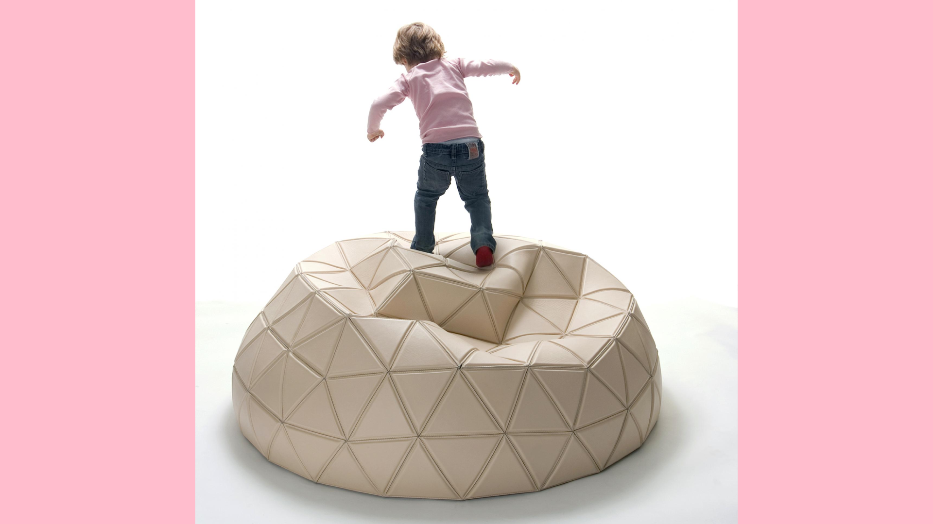 A young child is standing on a large, beige, geometrically-patterned bean bag, reminiscent of Buckminster Fuller's geodisic domes