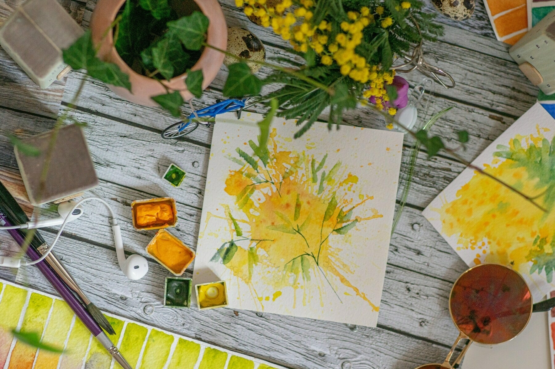A creative workspace with watercolor paintings featuring yellow and green floral motifs, paint cubes, brushes, and earphones on a rustic wooden table. Amongst these items are a potted plant, a bouquet of yellow wildflowers, and a pair of sunglasses, all suggesting an artist's break in progress.