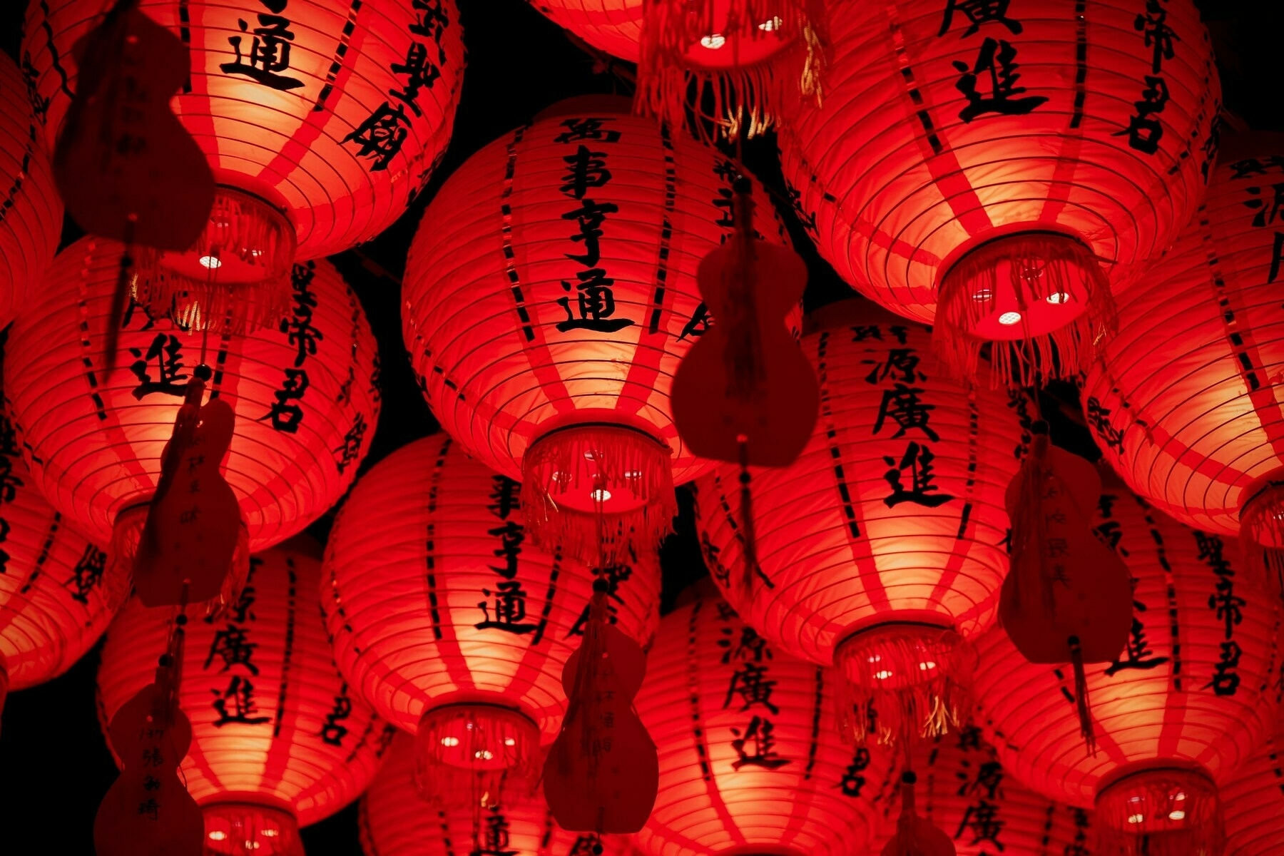 A collection of illuminated red Chinese lanterns with tassels and inscriptions, closely clustered against a dark background, creating a luminous and festive display.
