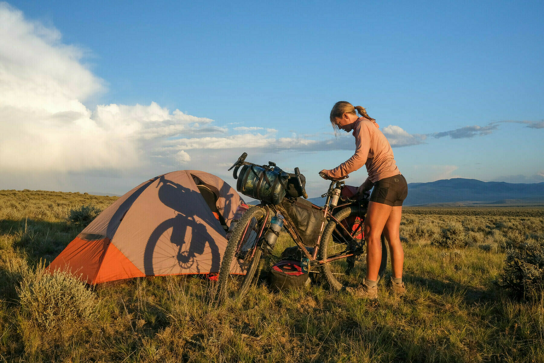 A camper adjusts their gear on a touring bicycle next to a tent with the shadow of another bike on it in an open field at sunset, with distant mountains and a cloudy sky in the background.