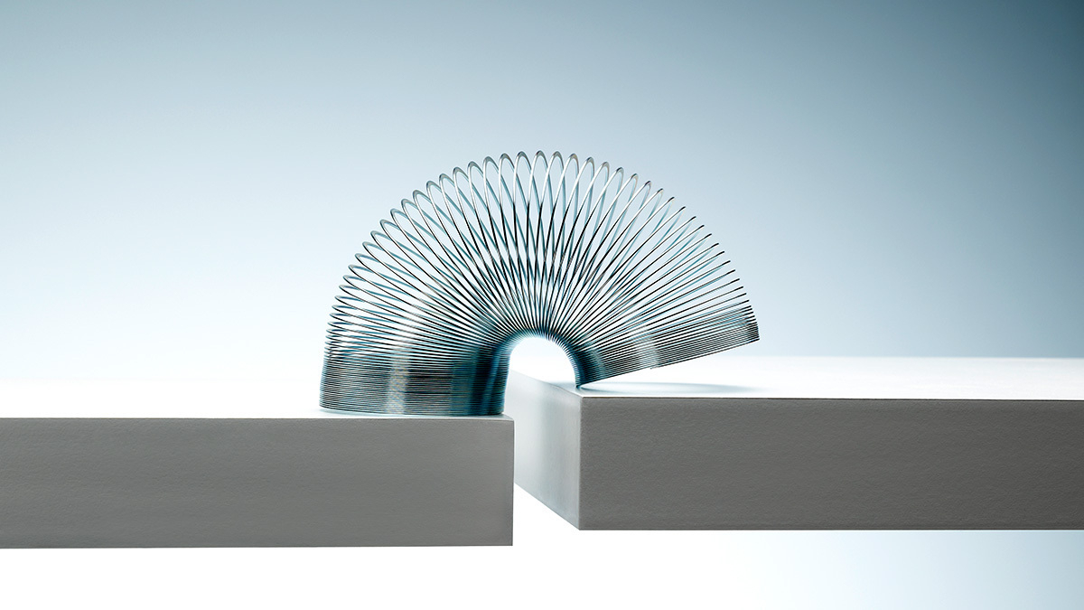 A metal slinky toy forms an arch between two white, matte surfaces under soft gradient lighting.