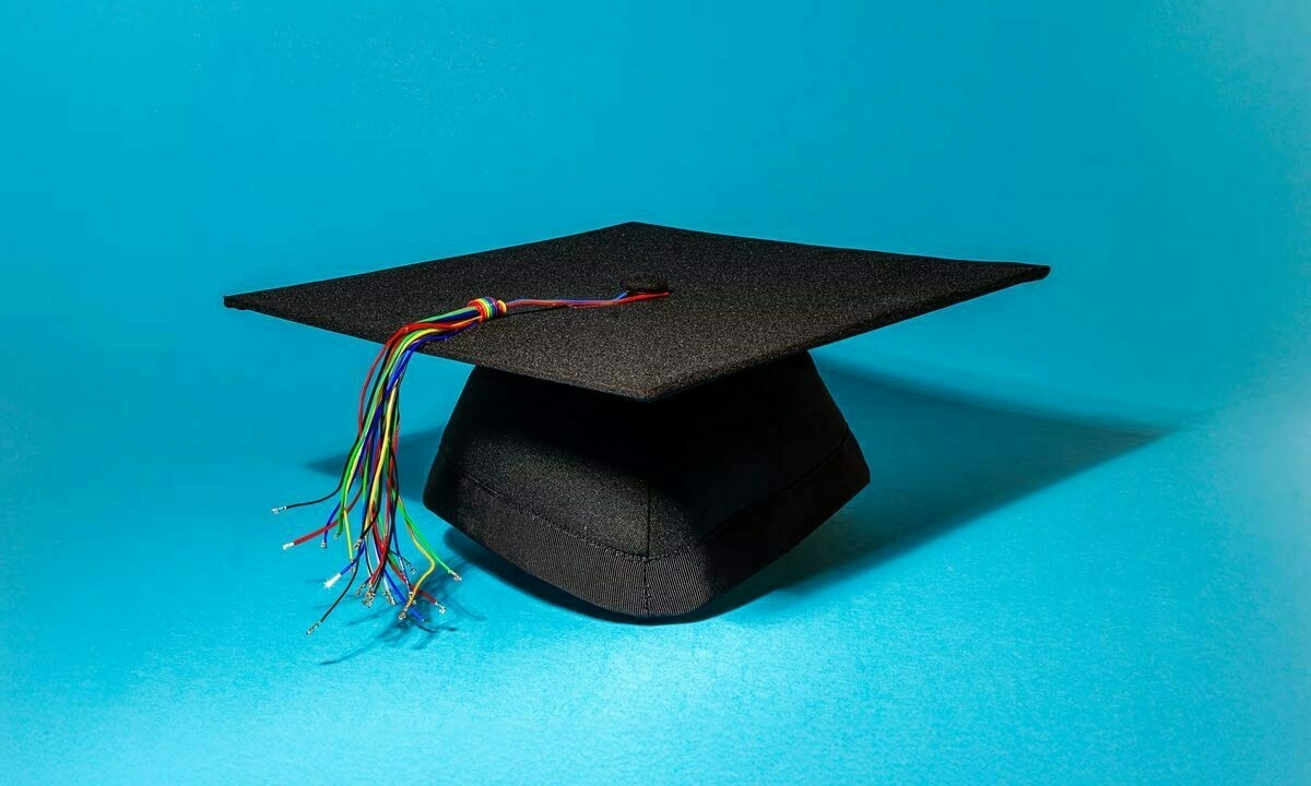 The image features a black graduation cap set against a bright blue background. The cap, traditionally square-shaped with a small button at the top center, is positioned to showcase its structure. Attached to the button is a tassel made of multicolored electrical wires, with several loose wire ends visible. The vibrant wires are in various colors, including red, yellow, green, and blue, creating a striking contrast against the solid black fabric and the smooth blue backdrop.