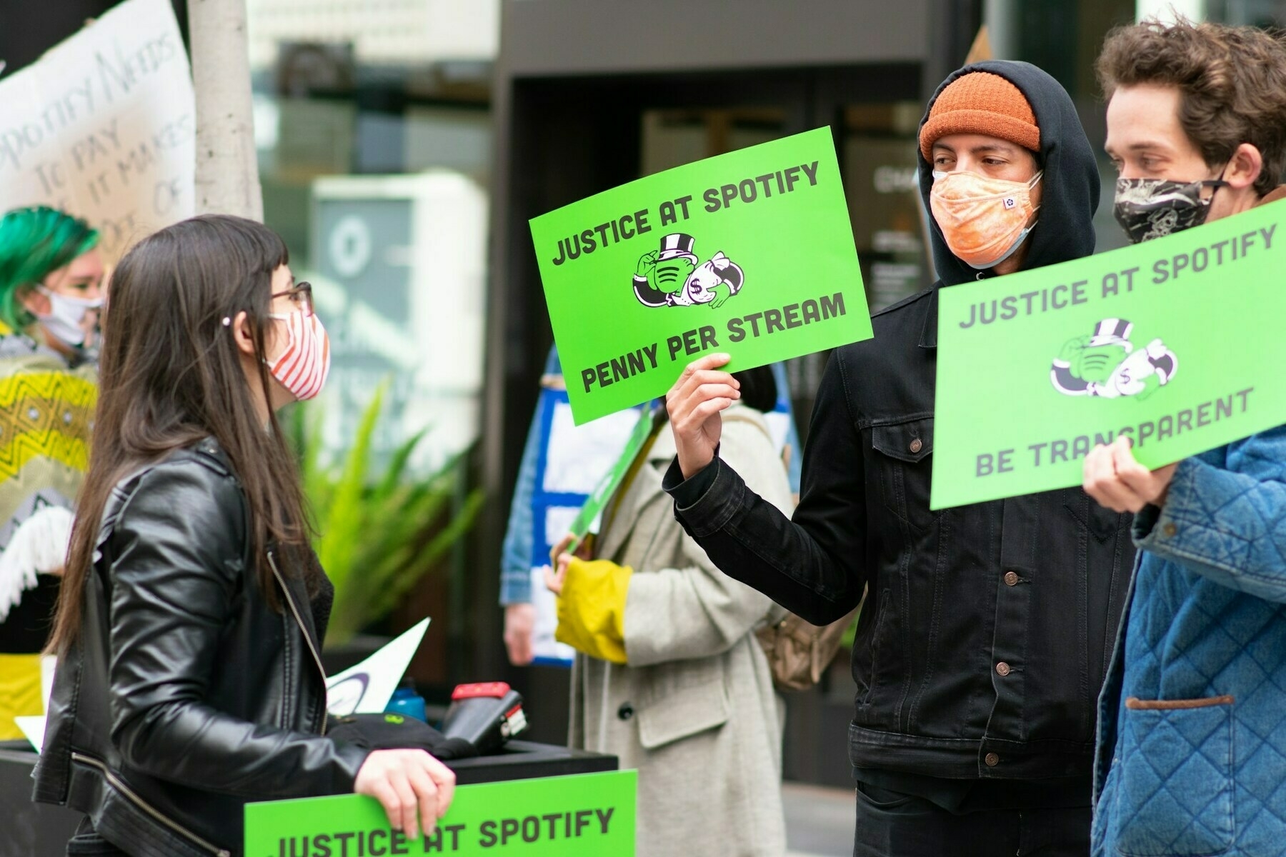 Union of Musicians and Allied Workers protesting at Spotify's corporate headquarters in San Francisco. A person in black jacket and face covering holds a sign demanding a penny per stream.