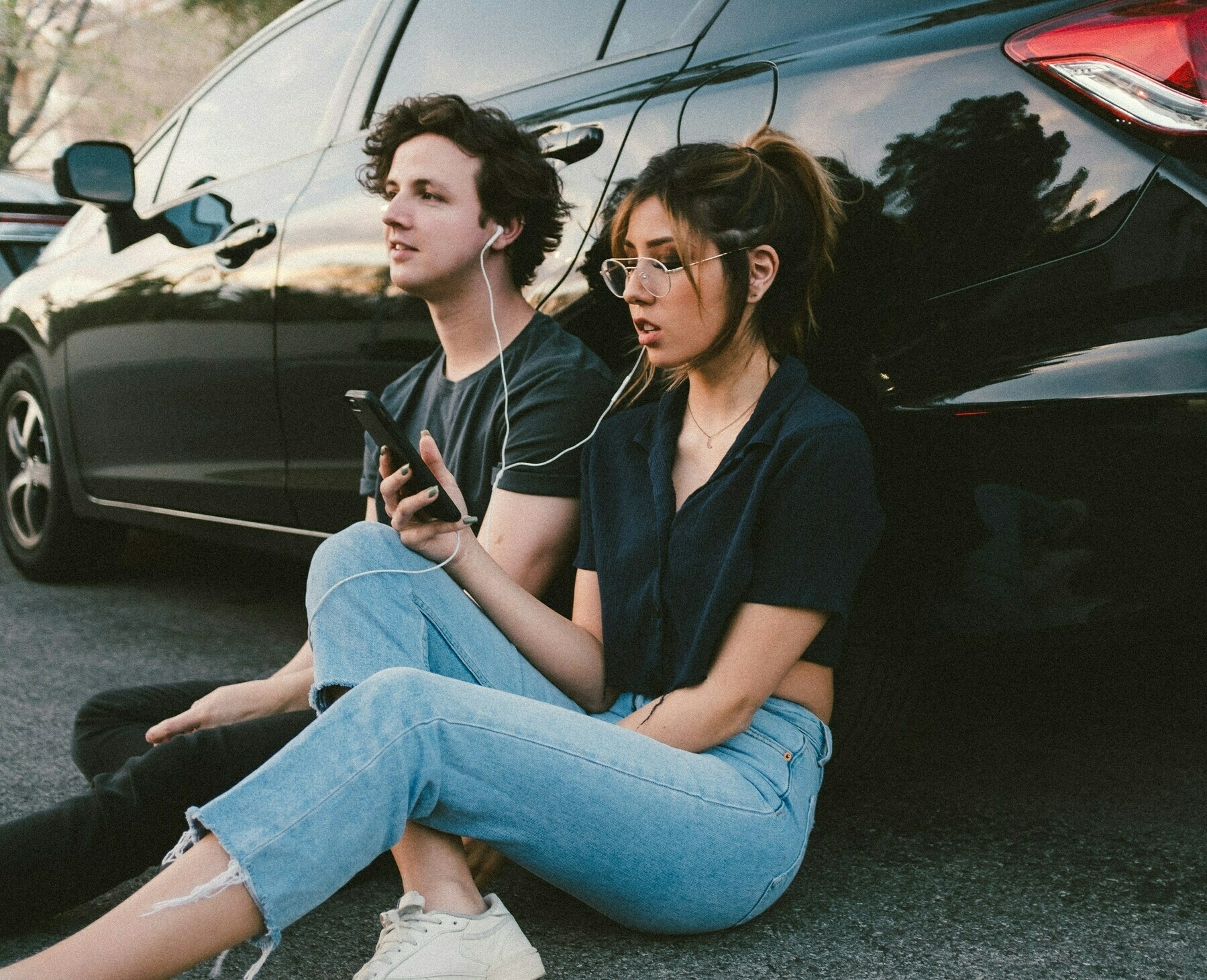 Two young people sitting on the ground with their backs to a car, sharing an earbud each 
