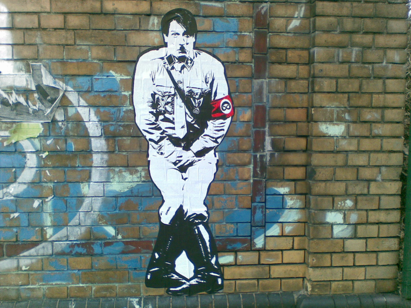 Auto-generated description: A street art piece depicts a stylized figure resembling a historical dictator with a red armband, set against a brick wall background with graffiti.