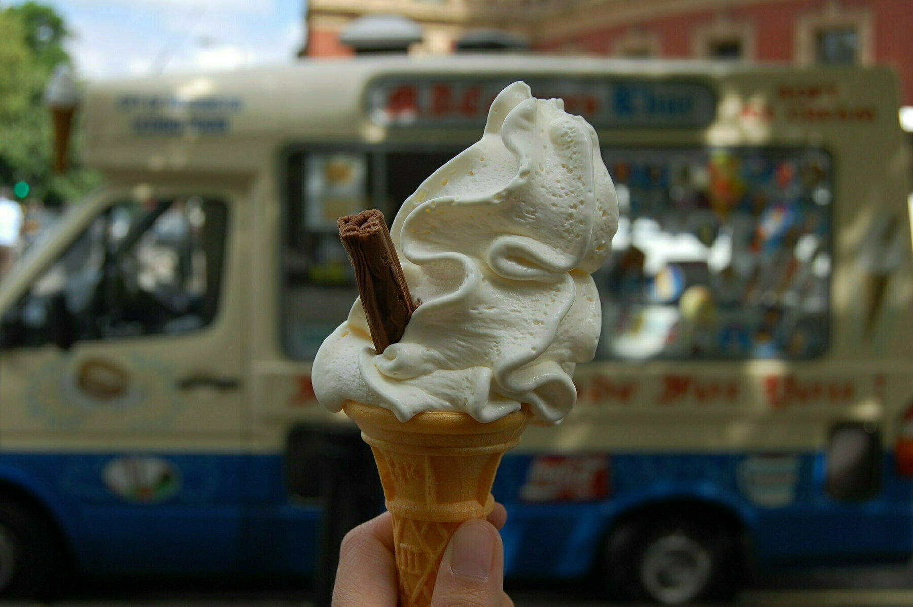 Auto-generated description: A hand is holding a soft-serve ice cream cone with a chocolate flake in front of an ice cream truck.