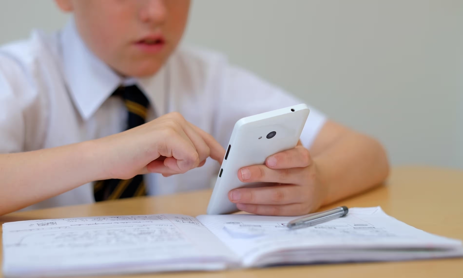 Child in uniform using a smartphone with an open notebook and pen on the table.