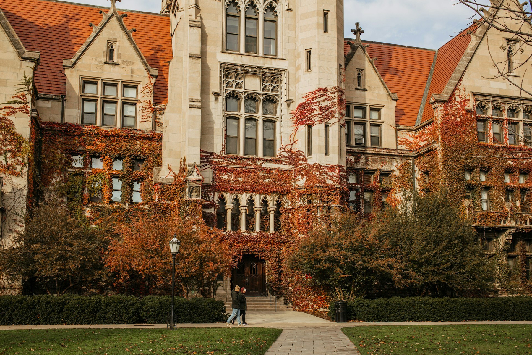 Auto-generated description: A historic building covered in autumn ivy with people walking along a path in front.