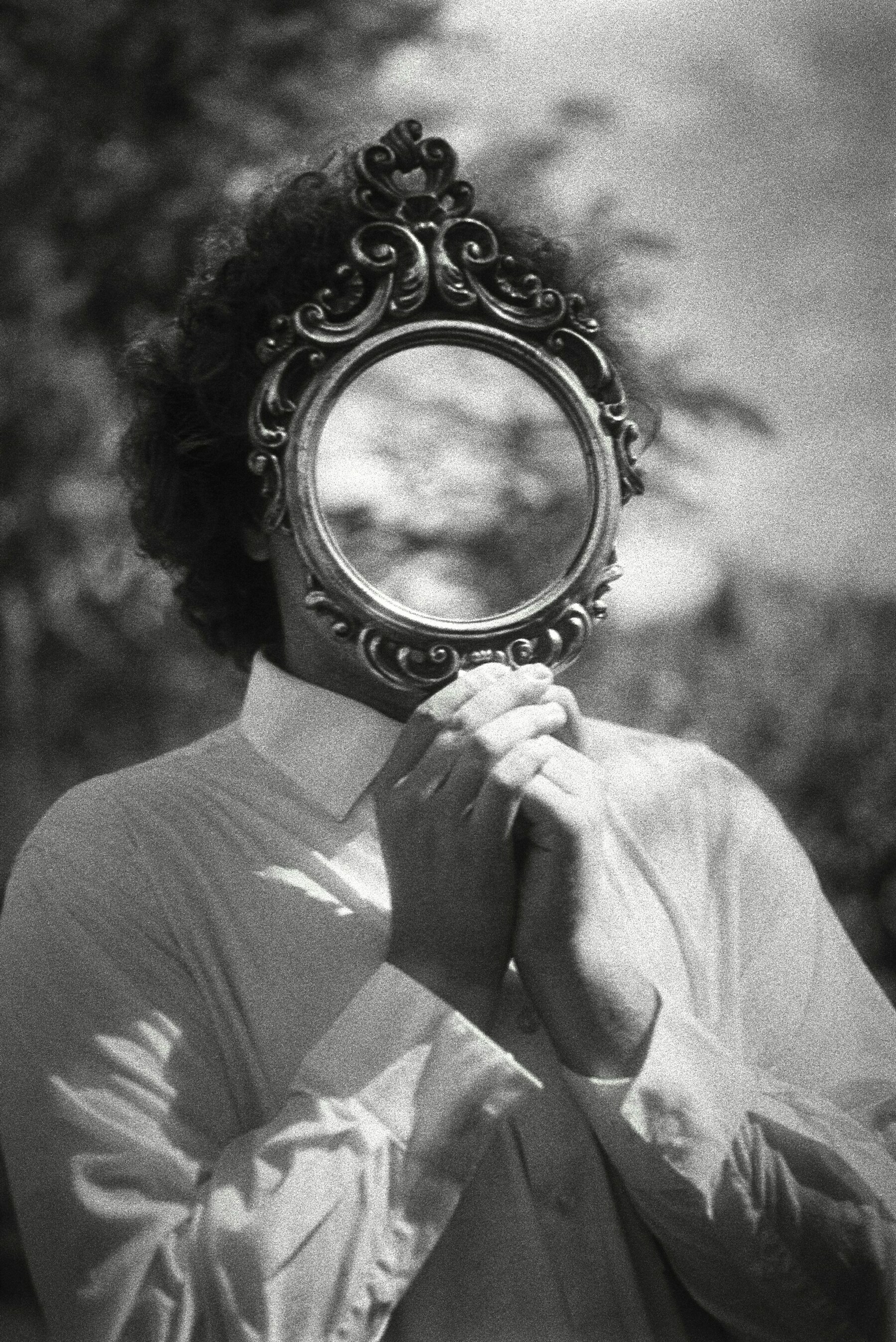 Auto-generated description: A person is holding a small ornate mirror in front of their face, creating a surreal effect as they wear a white shirt outdoors.