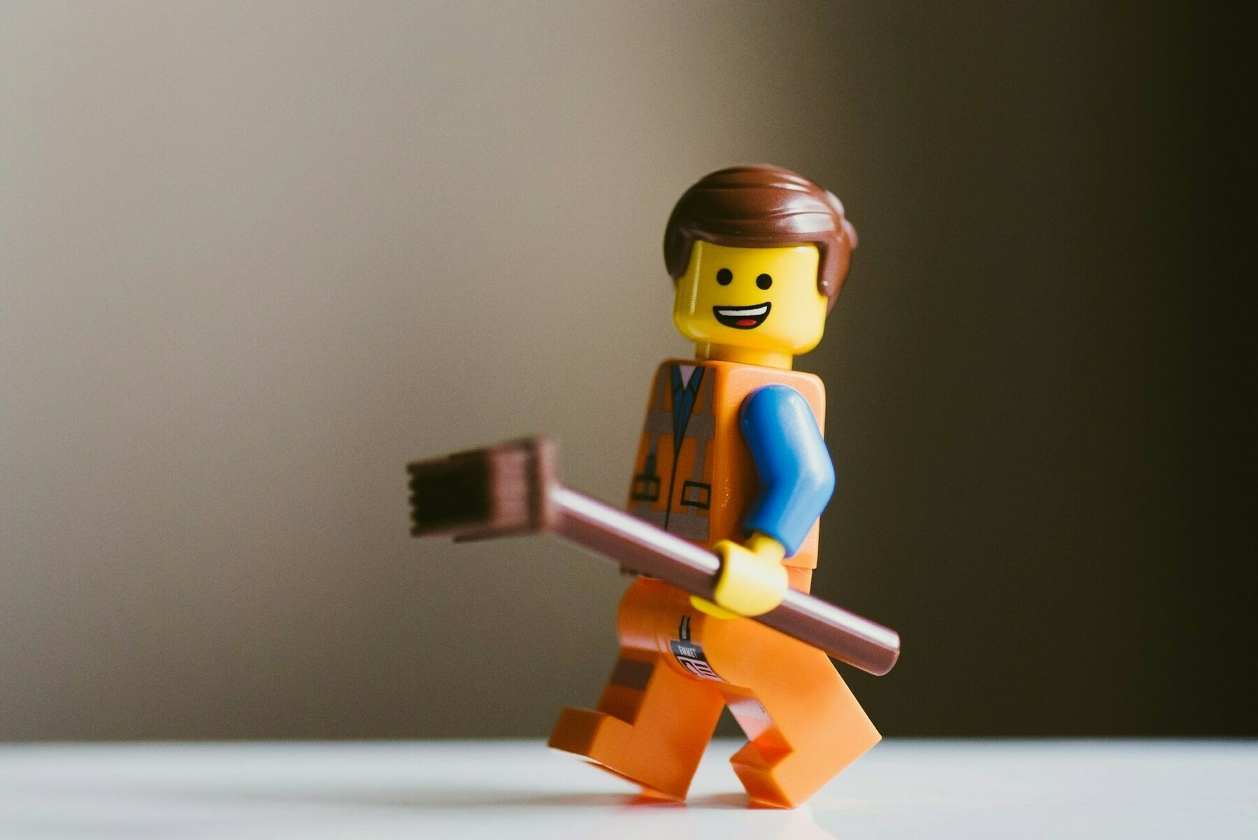 Auto-generated description: A Lego figure wearing an orange vest is holding a brown broom and smiling.
