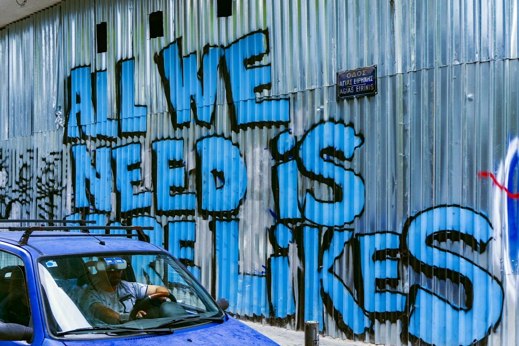 Auto-generated description: Graffiti on a corrugated metal wall reads ALL WE NEED IS LIKES in large blue letters, while a person drives a blue car nearby.