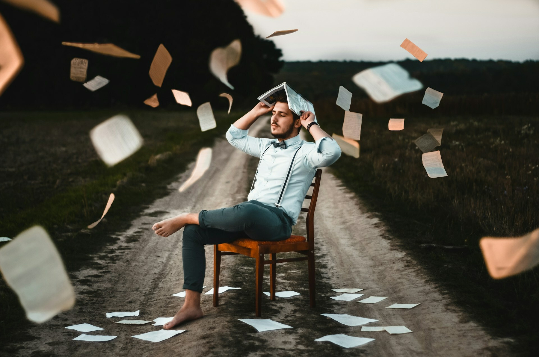 Auto-generated description: A man sitting on a chair in the middle of a dirt road is surrounded by floating sheets of paper, with one sheet used as a hat on his head.