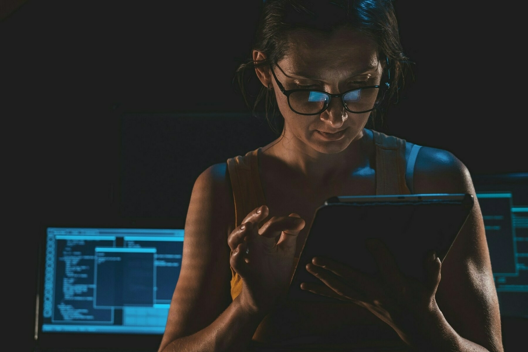 Auto-generated description: A person, wearing glasses, is focused on a tablet with two monitors in the background.