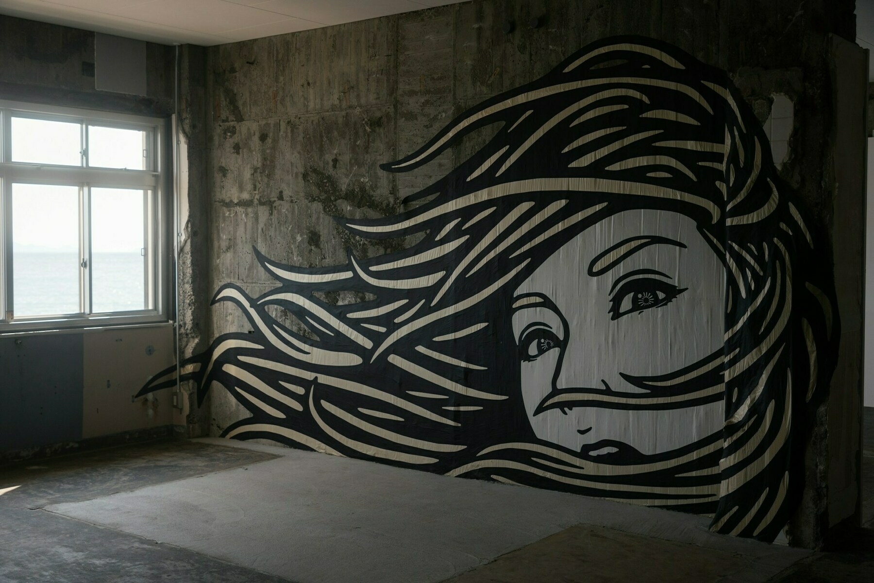 Auto-generated description: A mural of a woman's face with flowing hair covers a wall in an unfinished room with a window.