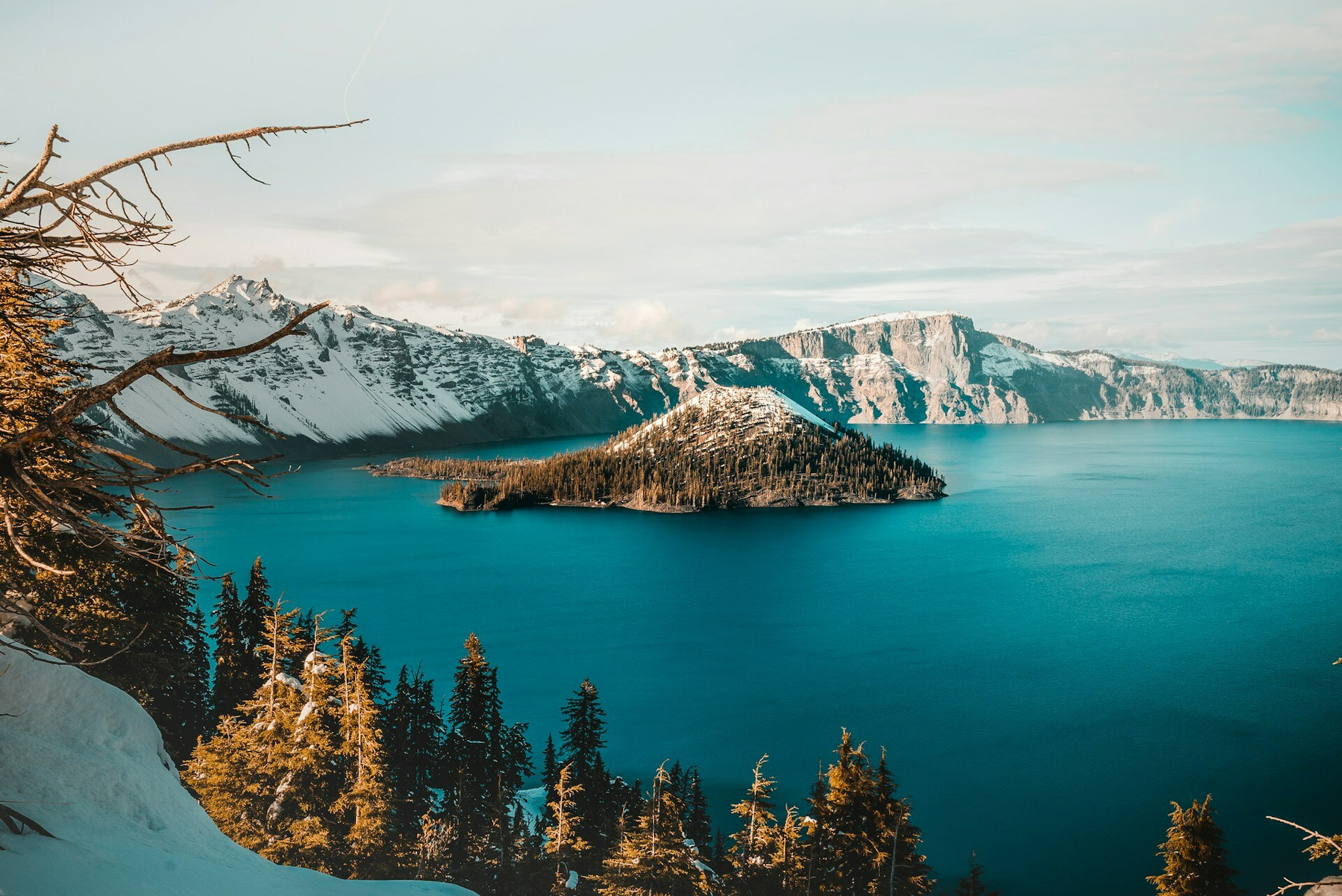 Auto-generated description: A snow-capped mountain range surrounds a blue crater lake with an island, framed by evergreen trees in the foreground.