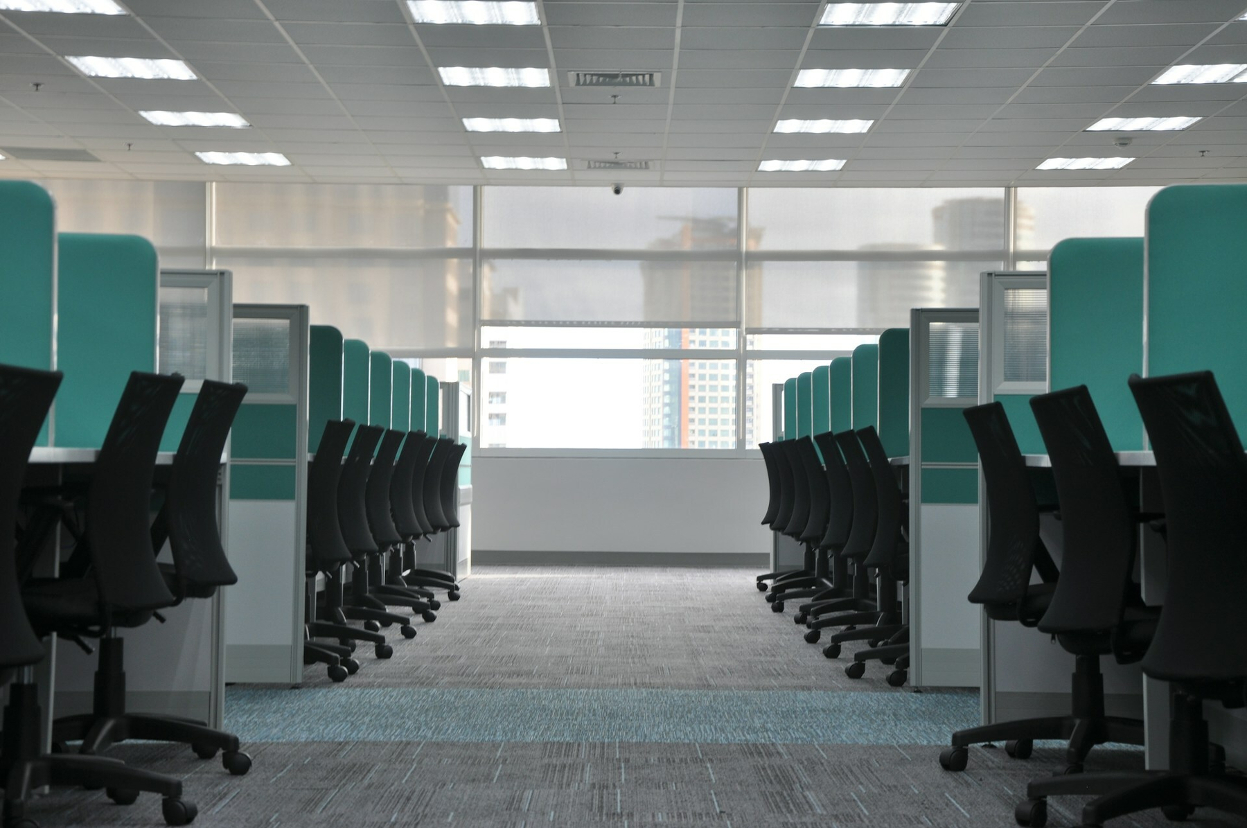 Auto-generated description: A modern office interior features rows of empty workstations with black chairs and blue partitions, illuminated by ceiling lights, with a large window at the end.