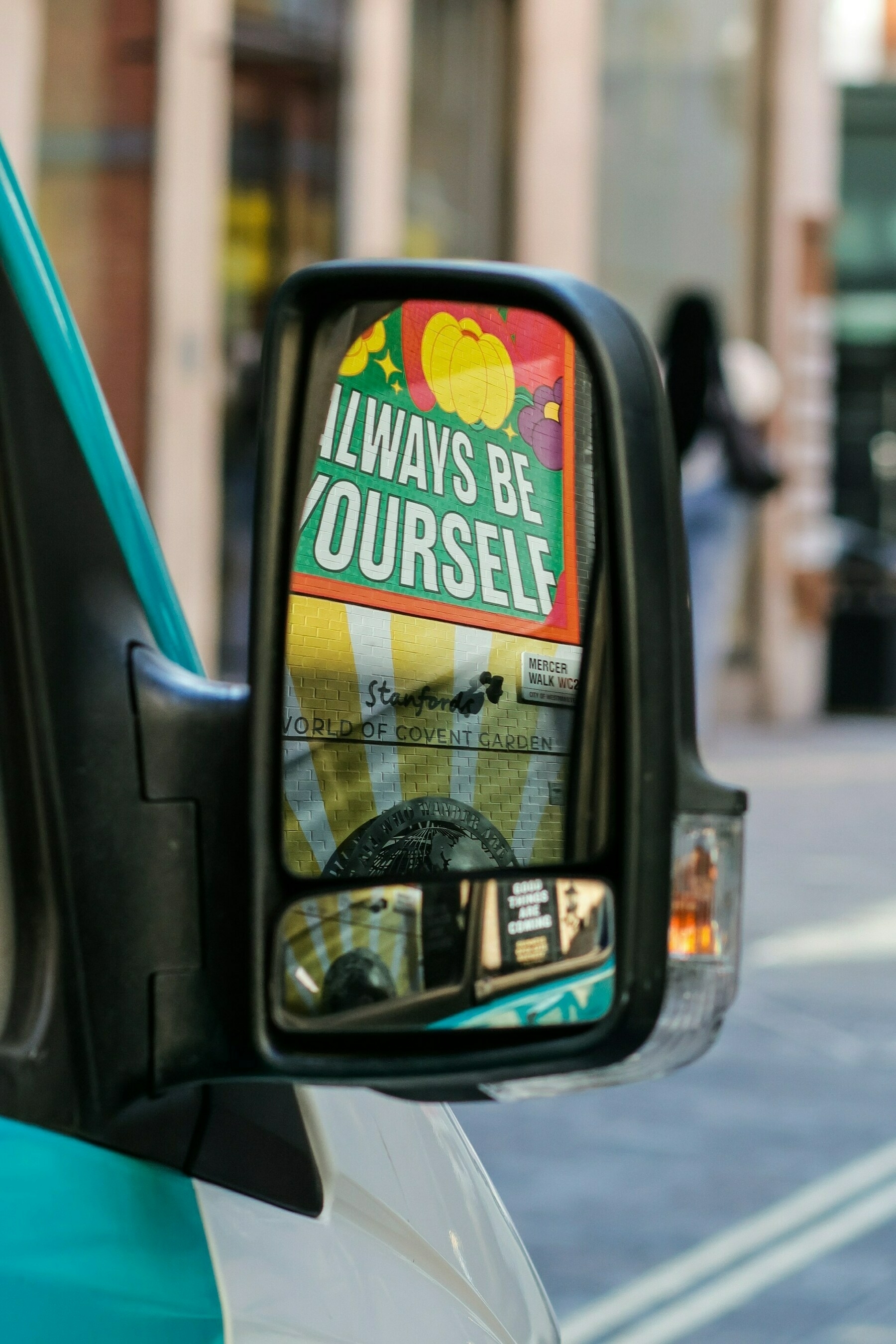Auto-generated description: A side mirror on a vehicle reflects a colorful sign saying ALWAYS BE YOURSELF along a street scene.