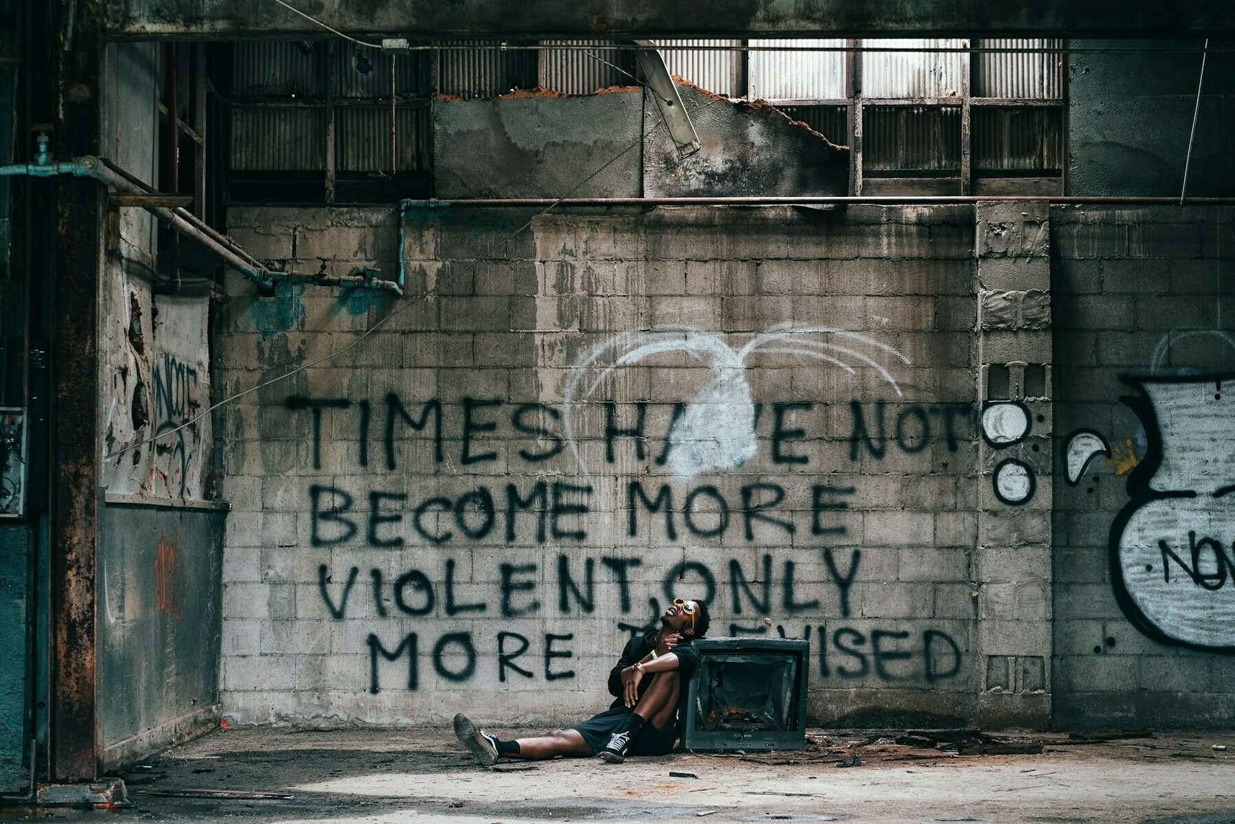 Auto-generated description: A person is sitting and resting in an abandoned industrial building with graffiti on the wall that reads, TIMES HAVE NOT BECOME MORE VIOLENT ONLY MORE TELEVISED.