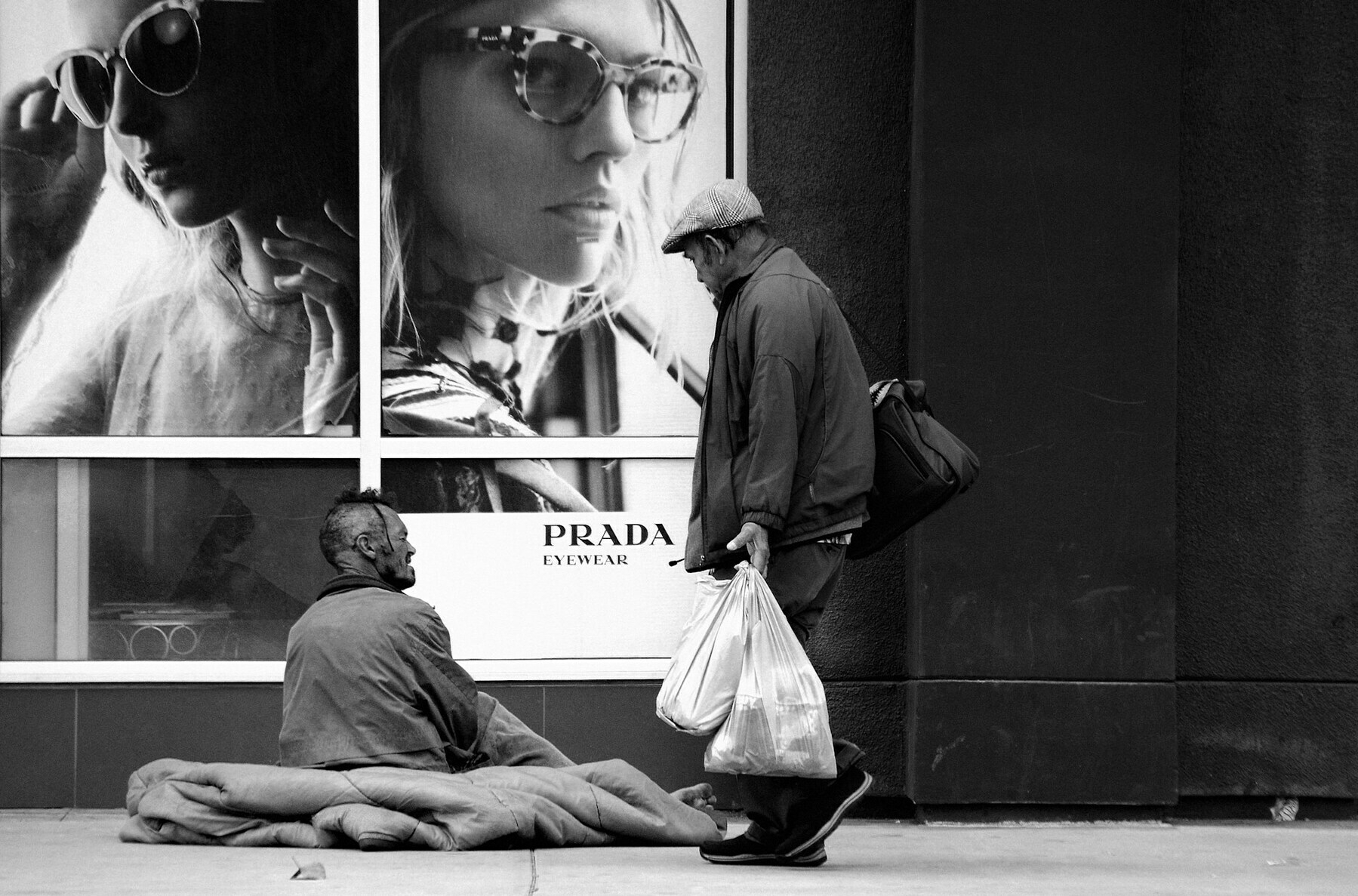 Grayscale photo of man carrying bags of shpping while walking past a male homeless person sitting on pavement outside a Prada store