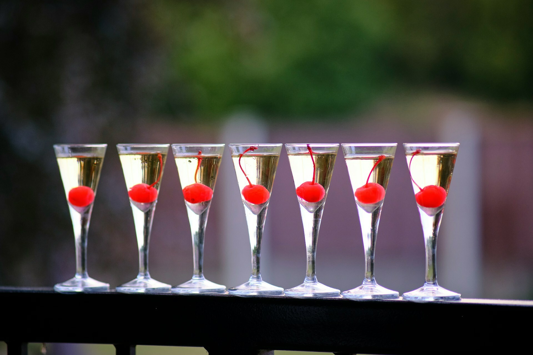 Auto-generated description: Six champagne glasses filled with a clear liquid and garnished with red cherries are lined up on a railing.