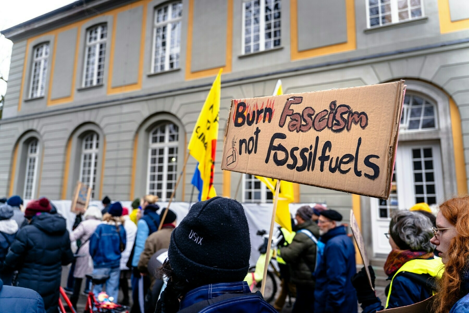 A group of people standing in front of a building, one is holding a sign that reads 'Burn Fascism not Fossil Fuels'