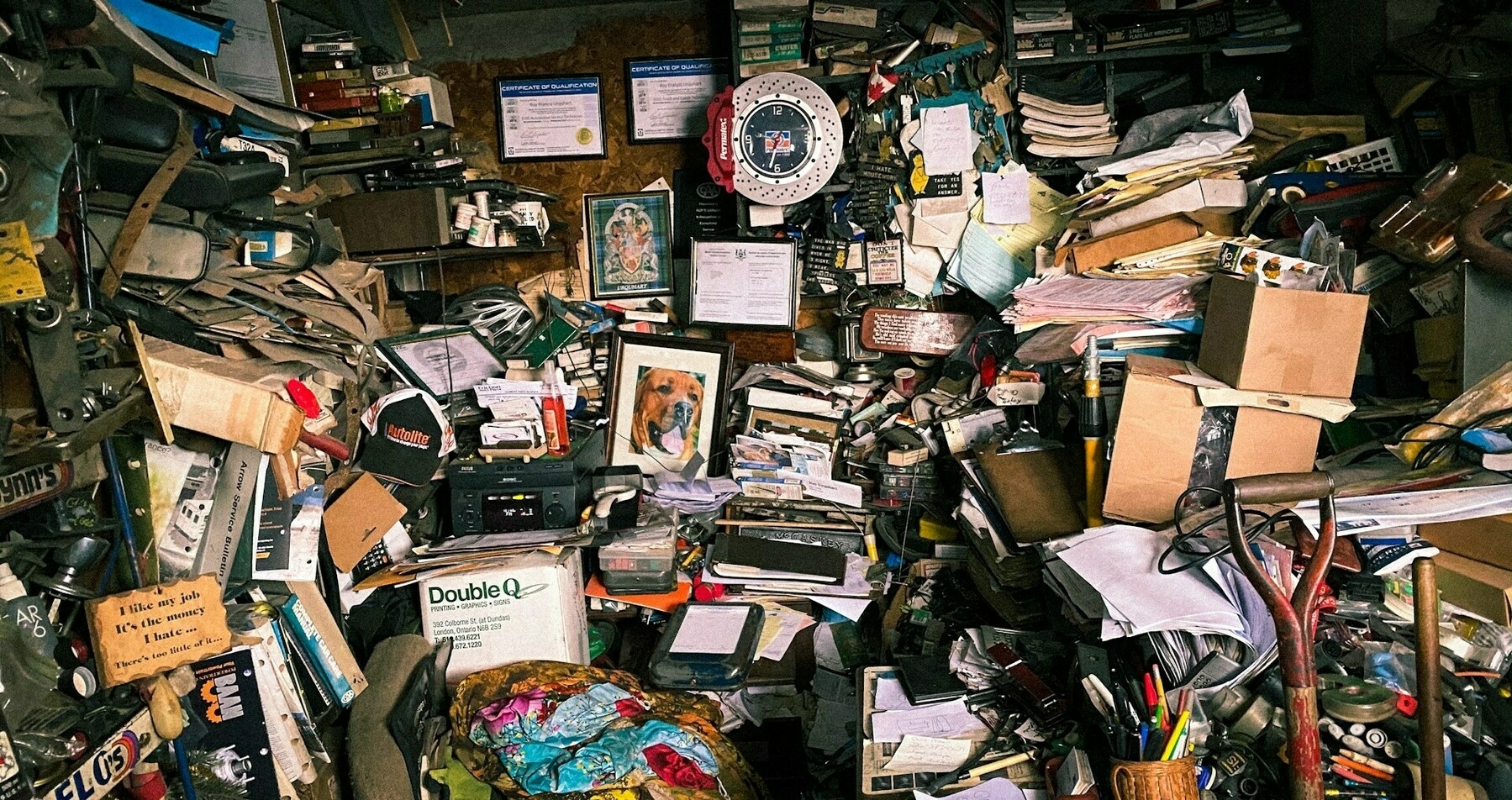 Auto-generated description: A cluttered room filled with stacks of papers, books, boxes, and various items, with certificates and a framed image on the wall.