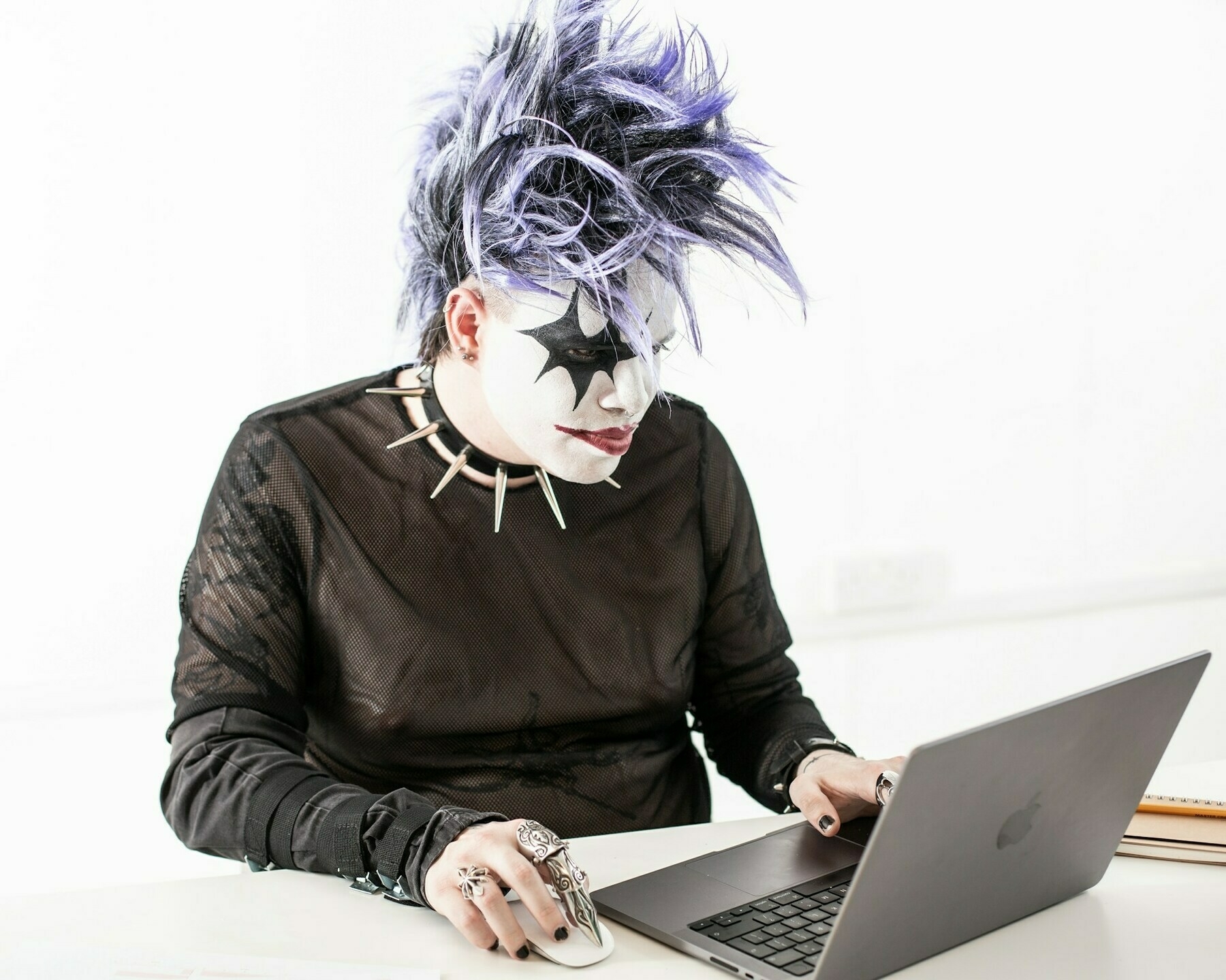 Person dressed in leather with face paint, a studded neck collar, and purple hair, using a laptop