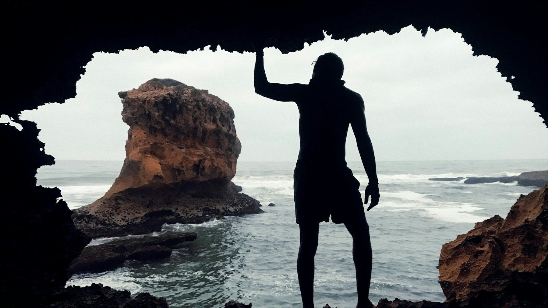 Auto-generated description: A person stands in silhouette inside a rocky cave, looking out at a large rock formation in the ocean.