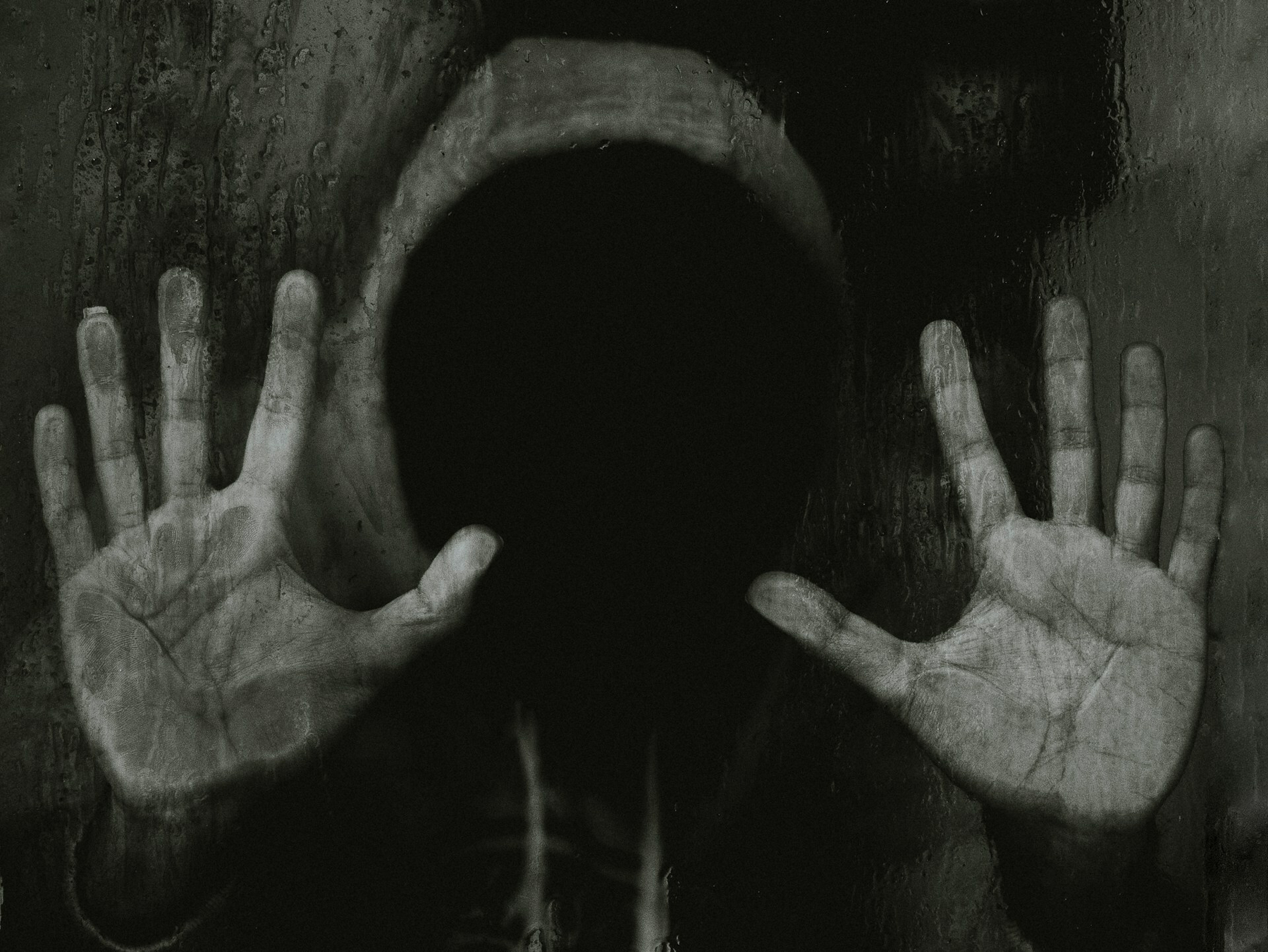 Auto-generated description: A person wearing a hood with an obscured face presses their hands against a dark, possibly wet, surface.