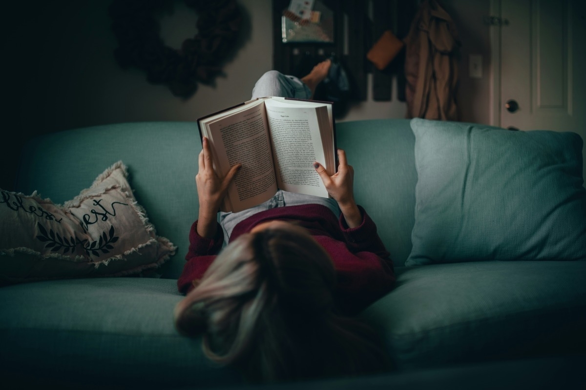 Person lying on a sofa reading a book in a dimly lit room.