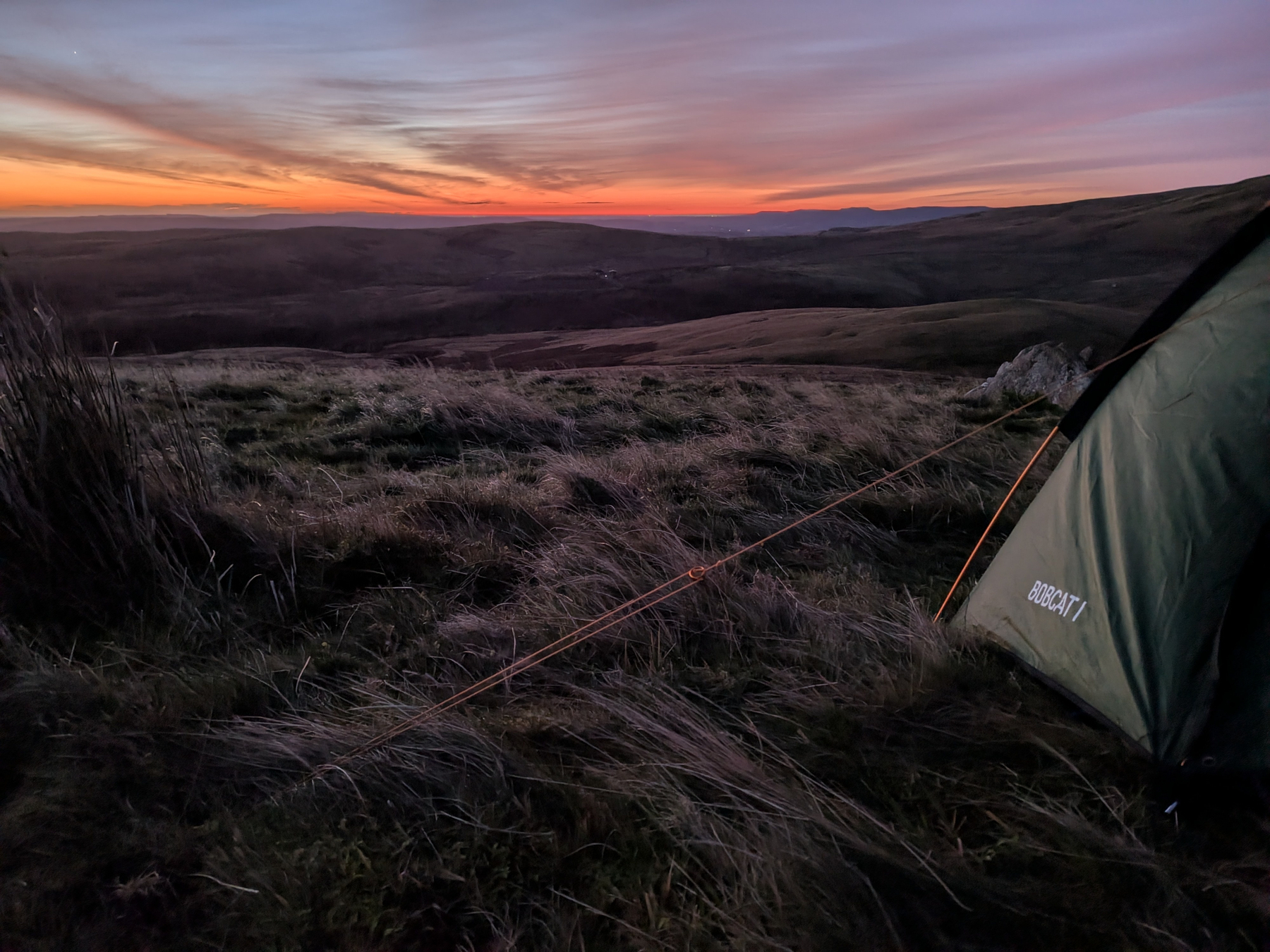 Auto-generated description: A tent is pitched on a grassy hillside during a colorful sunset, overlooking a vast landscape.