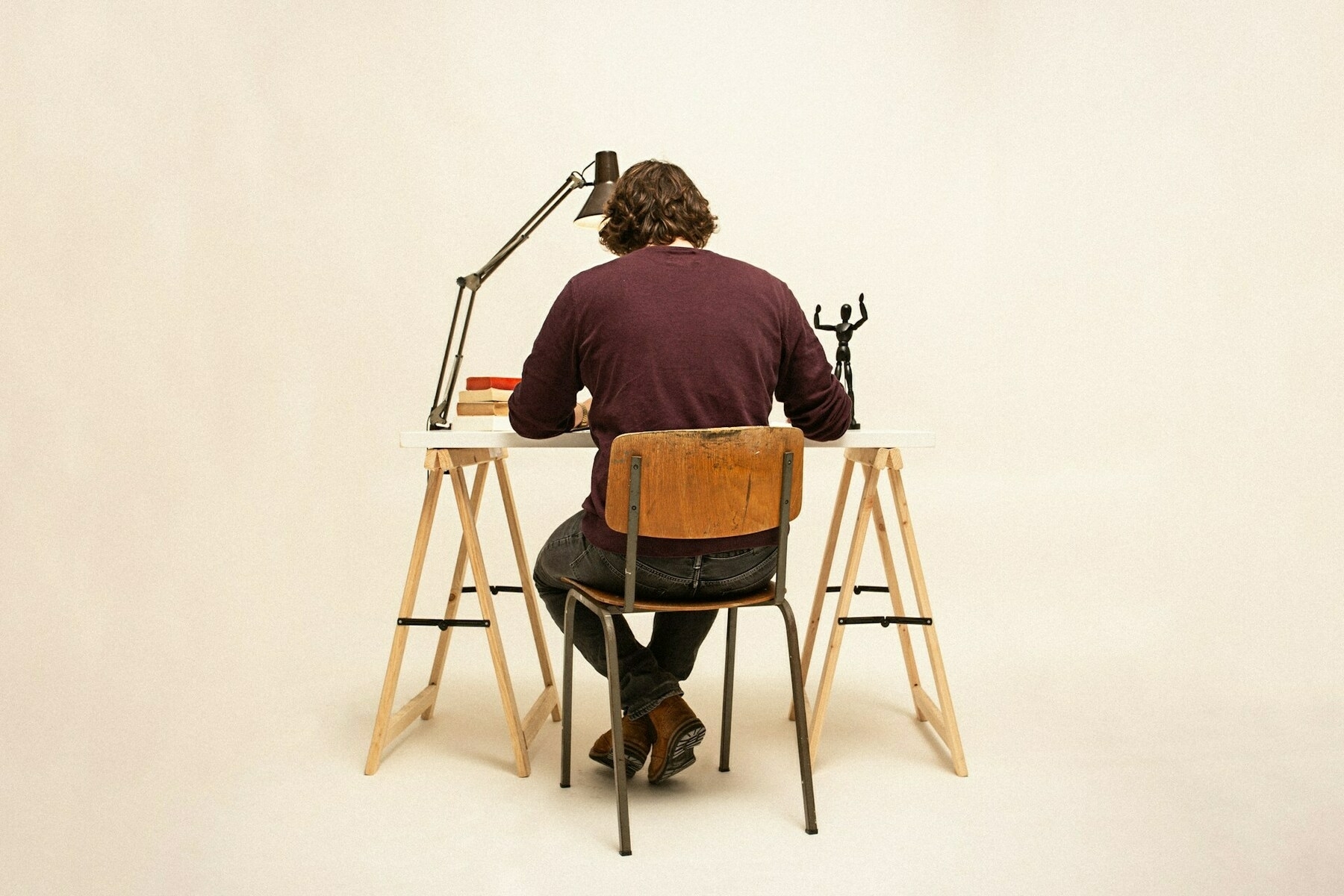 Auto-generated description: A person with long hair sits at a desk facing away, working under a desk lamp with books and decorative items nearby.