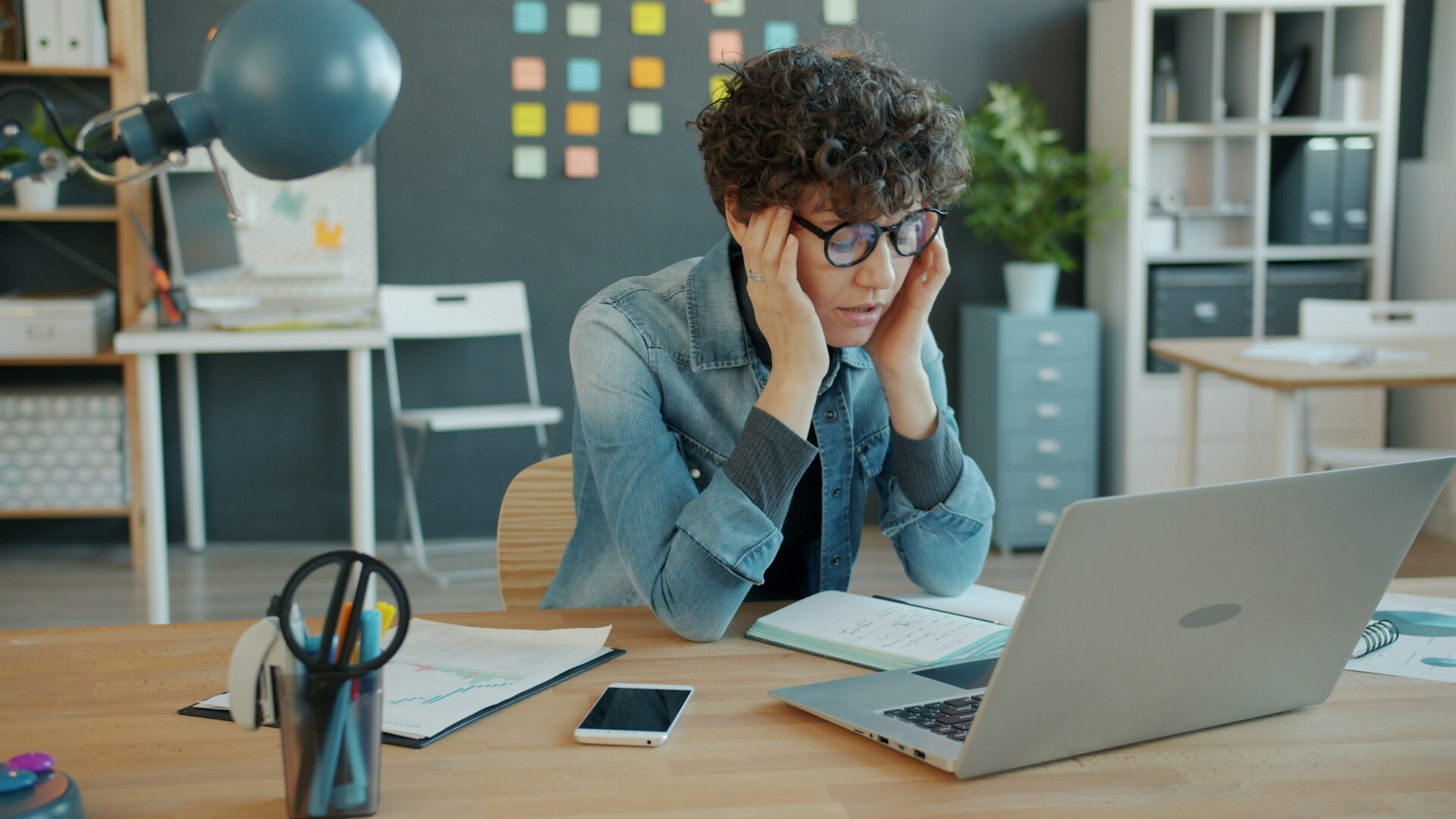 Auto-generated description: A person with curly hair and glasses appears to be focused or concerned while looking at a laptop in a modern office setting.