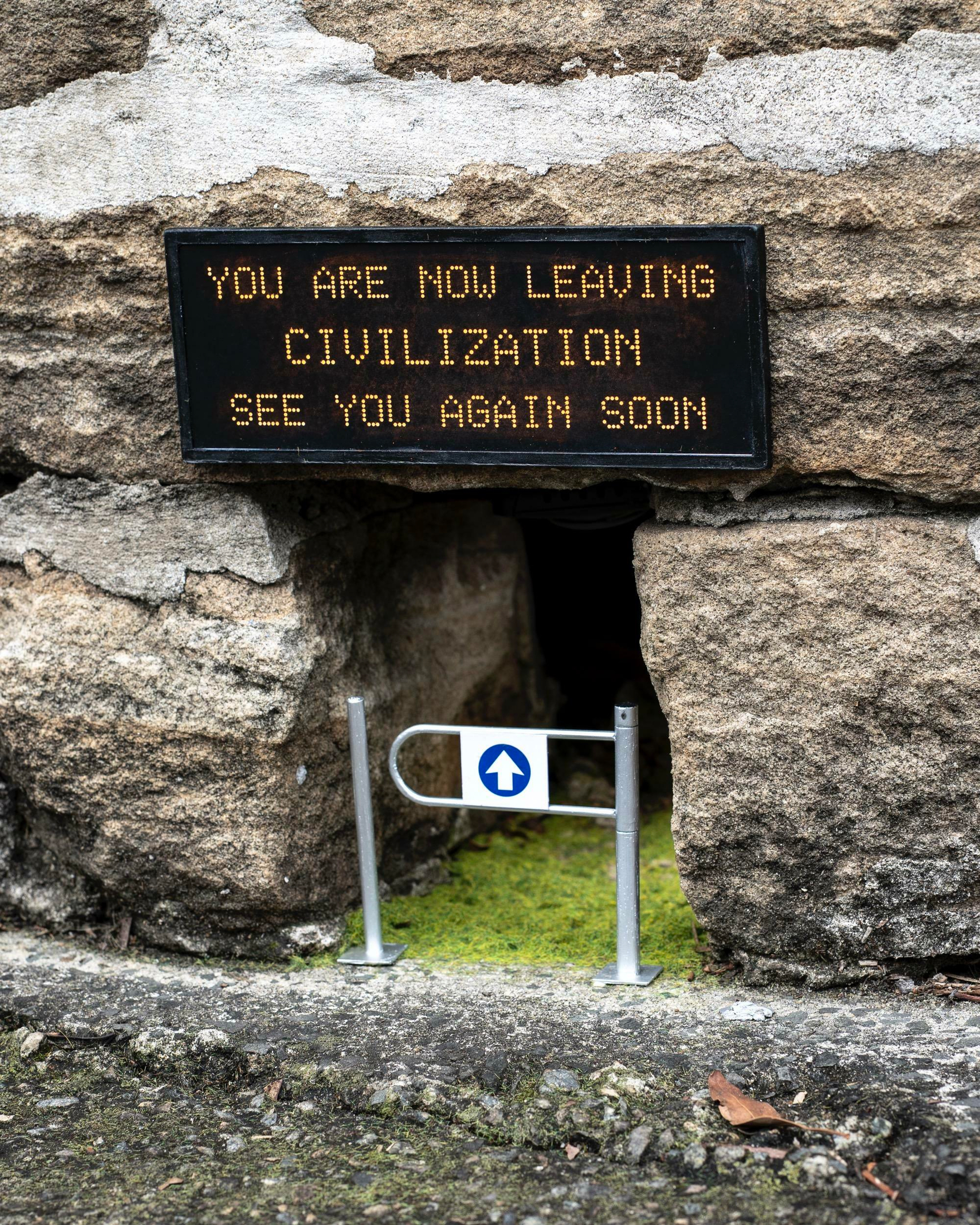 Auto-generated description: A small passageway through a stone wall features a humorous sign that reads YOU ARE NOW LEAVING CIVILIZATION, SEE YOU AGAIN SOON, accompanied by a tiny barrier and arrow.