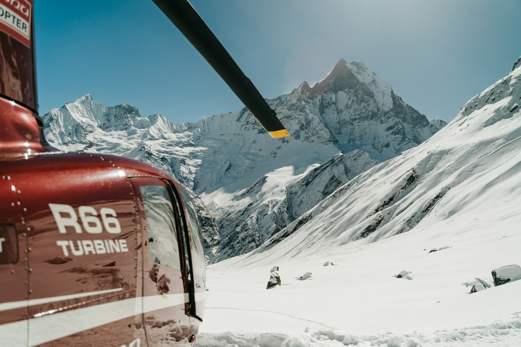 Auto-generated description: A red helicopter is parked on a snowy mountain slope with a backdrop of majestic, snow-covered peaks.