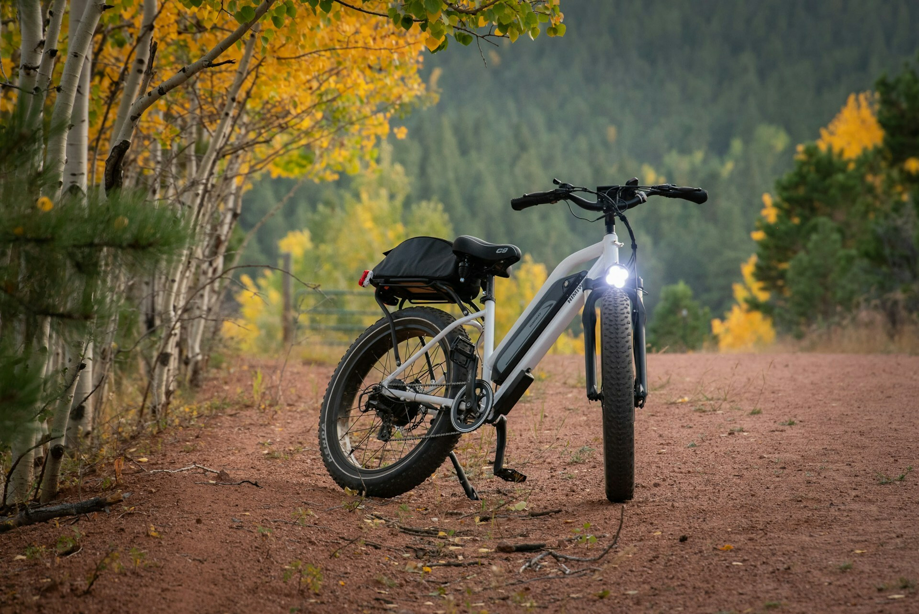 Auto-generated description: A mountain bike with large tires and a front light is parked on a dirt path surrounded by autumn trees.