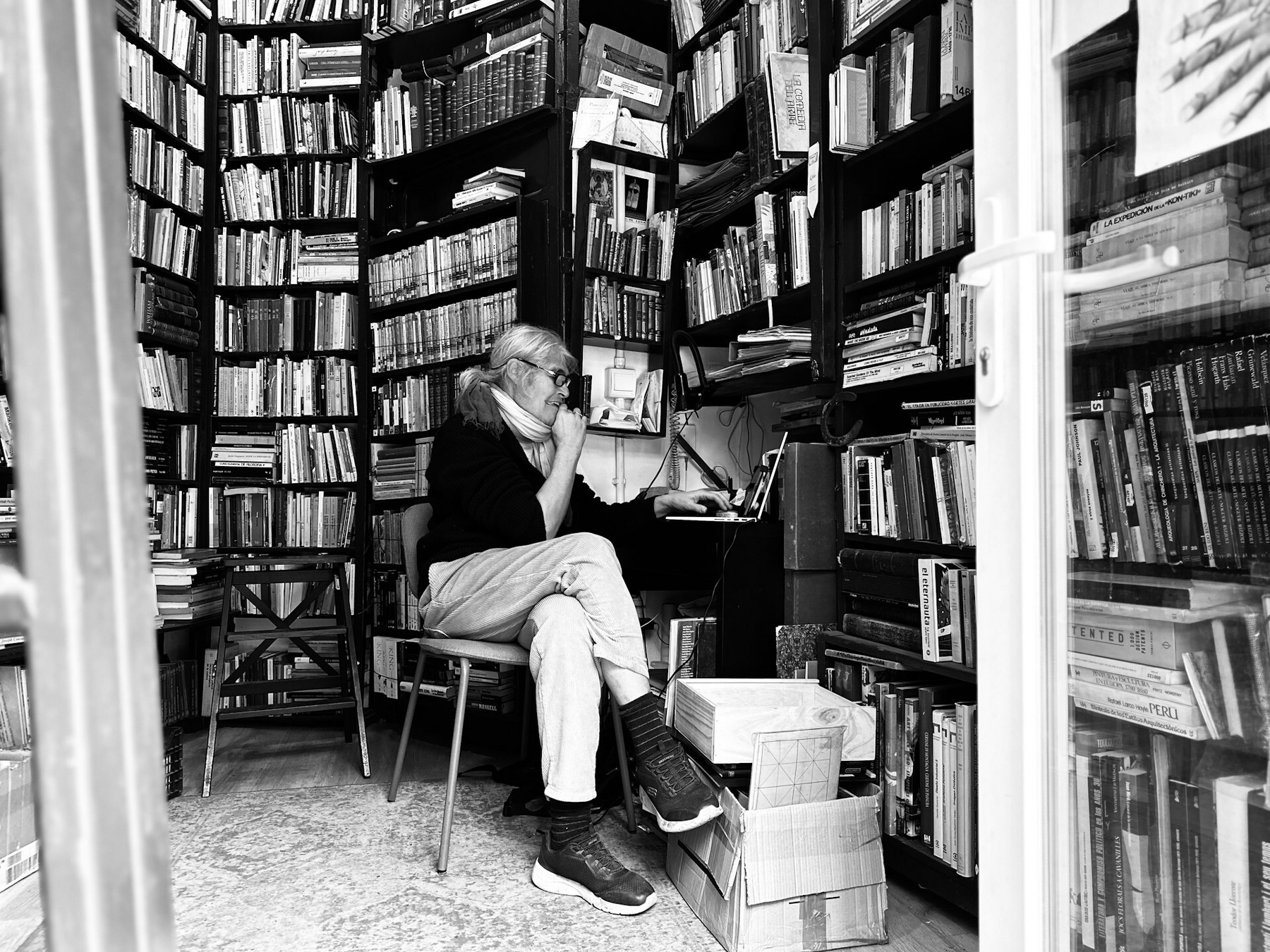Auto-generated description: A person sits in a small, book-filled room, engaged with a computer amidst towering shelves of books.