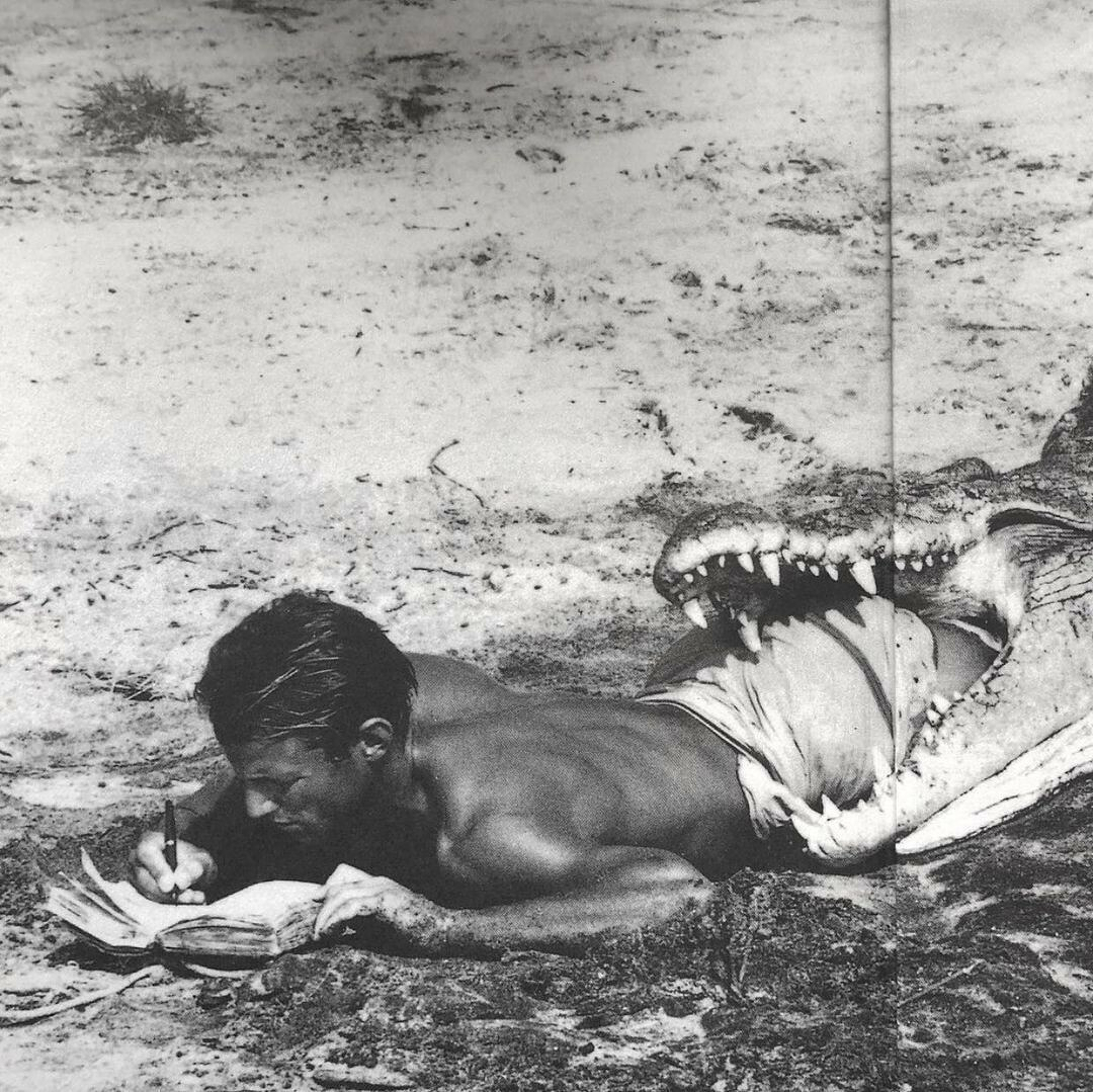 Auto-generated description: A man lies on the ground writing in a notebook while seemingly being swallowed by the open mouth of a large alligator or crocodile.