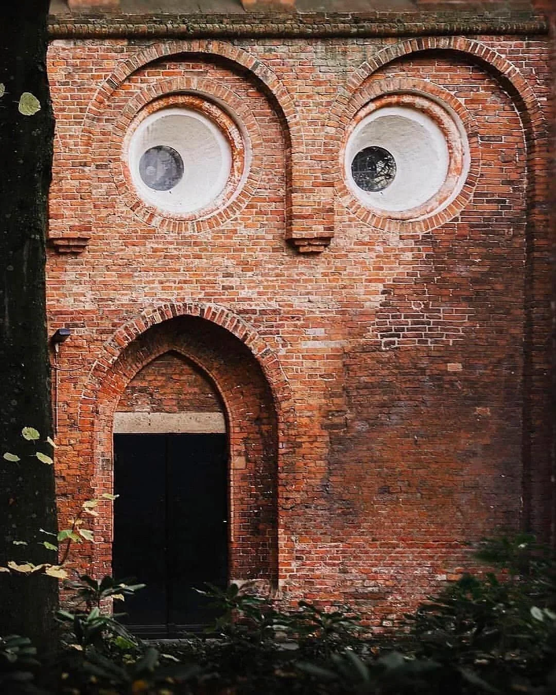 Auto-generated description: A brick wall with two circular windows and an arched doorway resembles a face.